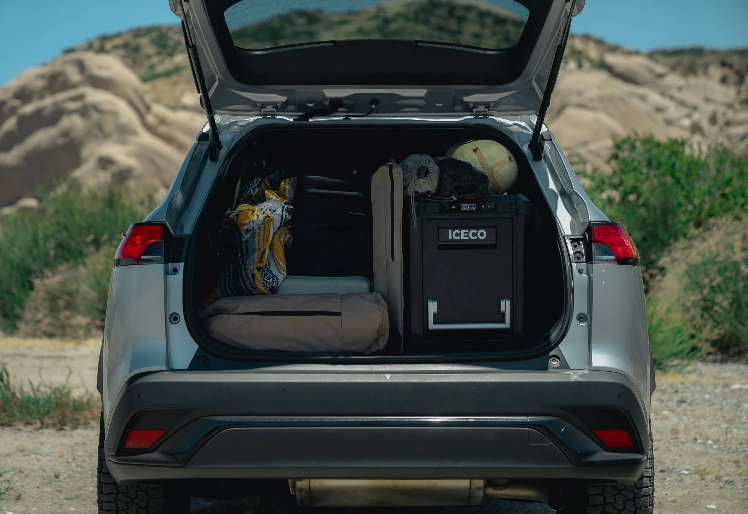 Open back of a silver SUV showing packed camping items inside, including a duffel bag, rolled-up sleeping mats, a helmet, and a yellow and black striped blanket, with a rocky landscape in the background.