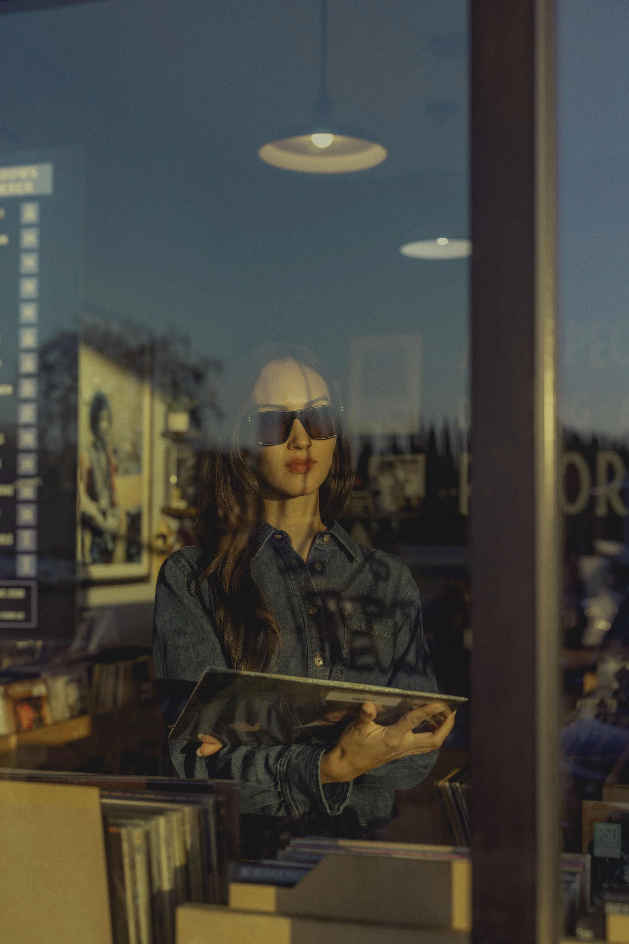 A woman with long wavy hair, wearing large dark sunglasses and a denim jacket, is standing inside a bookstore or library. She is holding and looking at a vinyl record album cover. The shop has shelves filled with books, and the scene is reflected in 