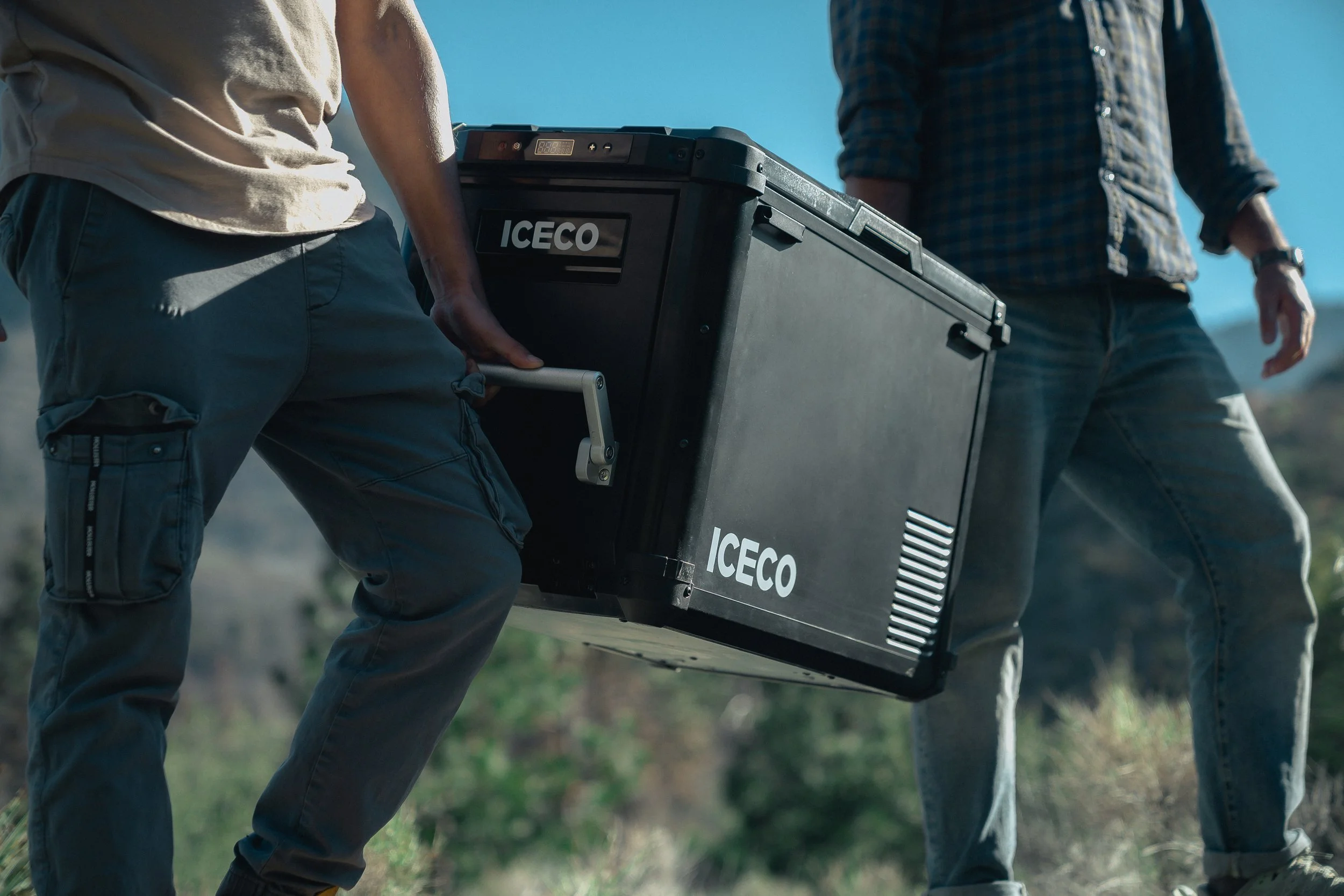 Two men outdoors lifting or carrying a black portable cooler with the brand name ICECO, under a clear blue sky.