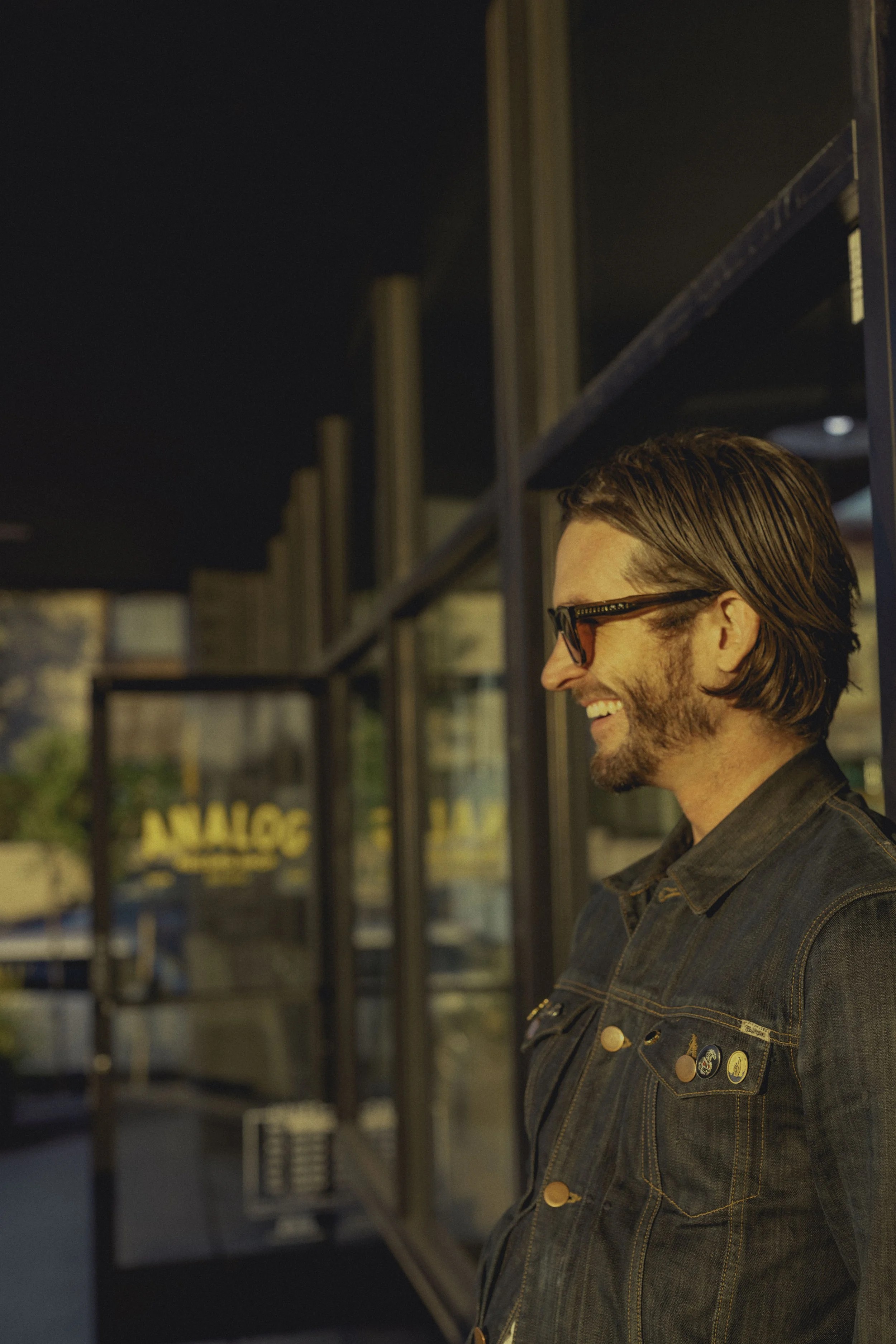 A man with brown hair, glasses, and a beard, wearing a denim jacket with pins, smiling and standing outdoors at dusk.
