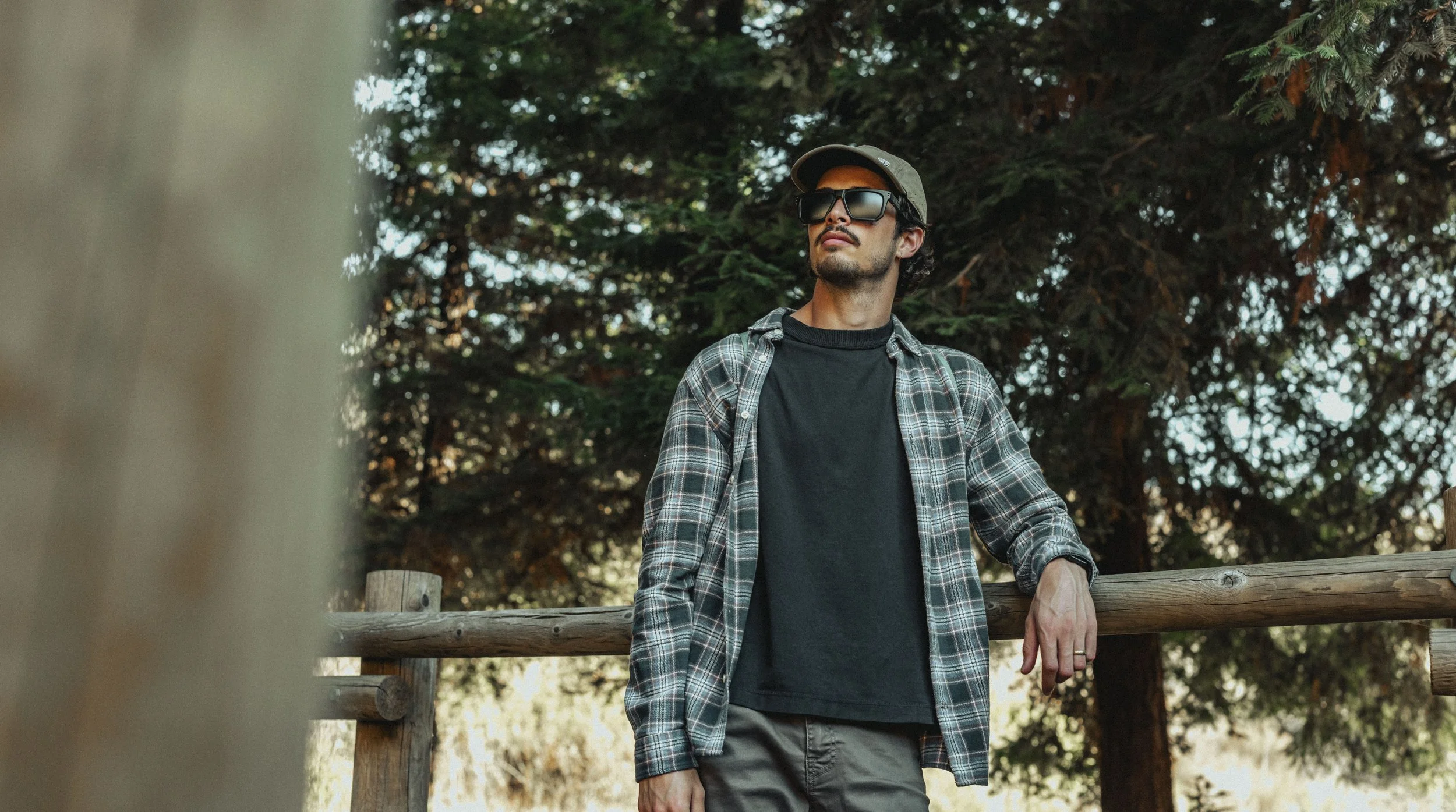 Young man wearing sunglasses, a cap, a flannel shirt, and a black t-shirt, leaning on a wooden railing outdoors surrounded by trees.
