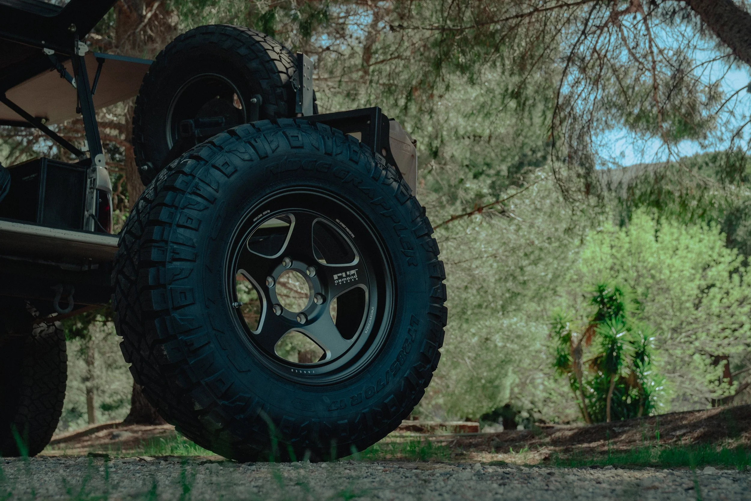 View of the rear tire and wheel of a vehicle on a dirt path, surrounded by trees and greenery.