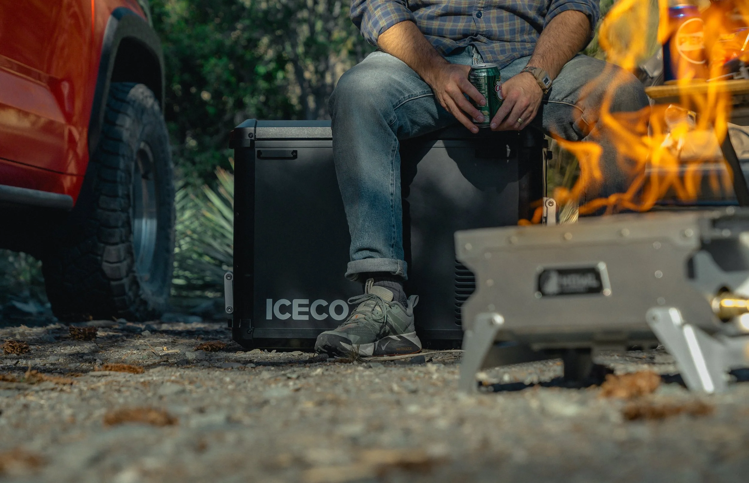 Person sitting outdoors near a grill, holding a soda can, with camping equipment and a vehicle nearby.