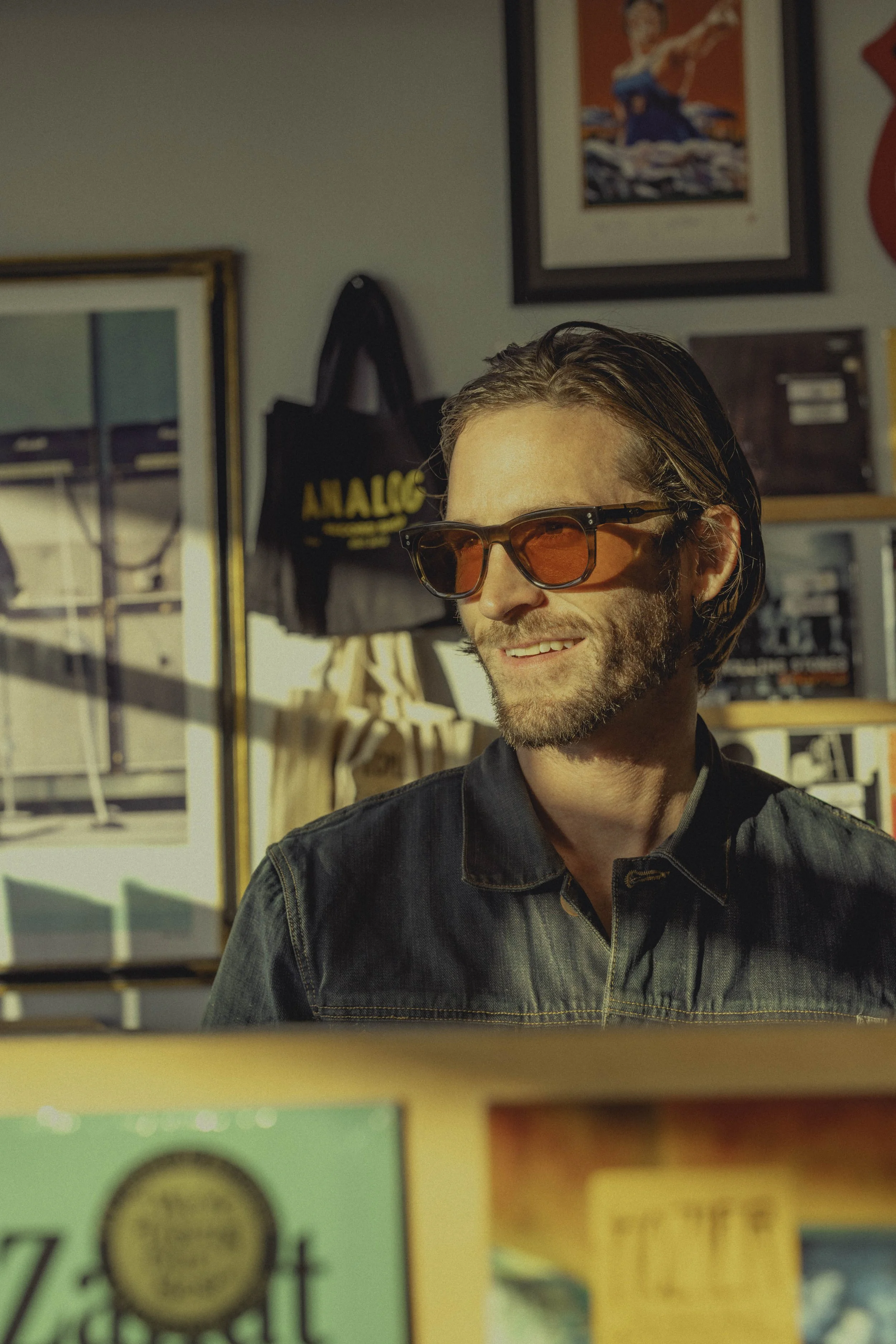 A man with shoulder-length brown hair,  glasses with orange lenses, and a beard, smiling and looking to the side, wearing a dark jacket. Behind him are framed artworks and a black tote bag with yellow text that says "AALOG".