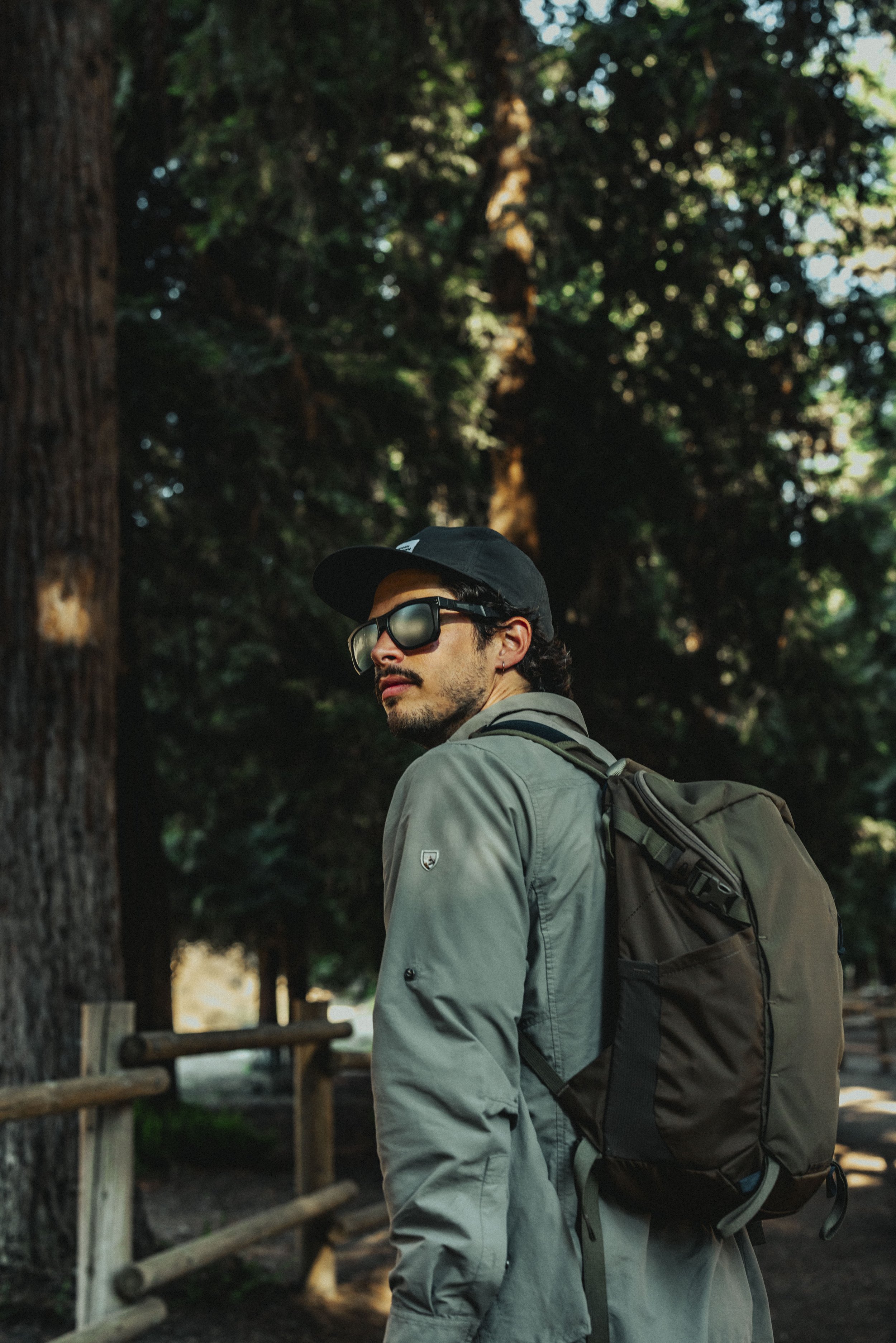 A man with a beard, wearing sunglasses, a black cap, a gray jacket, and carrying a backpack, standing outdoors among tall trees.