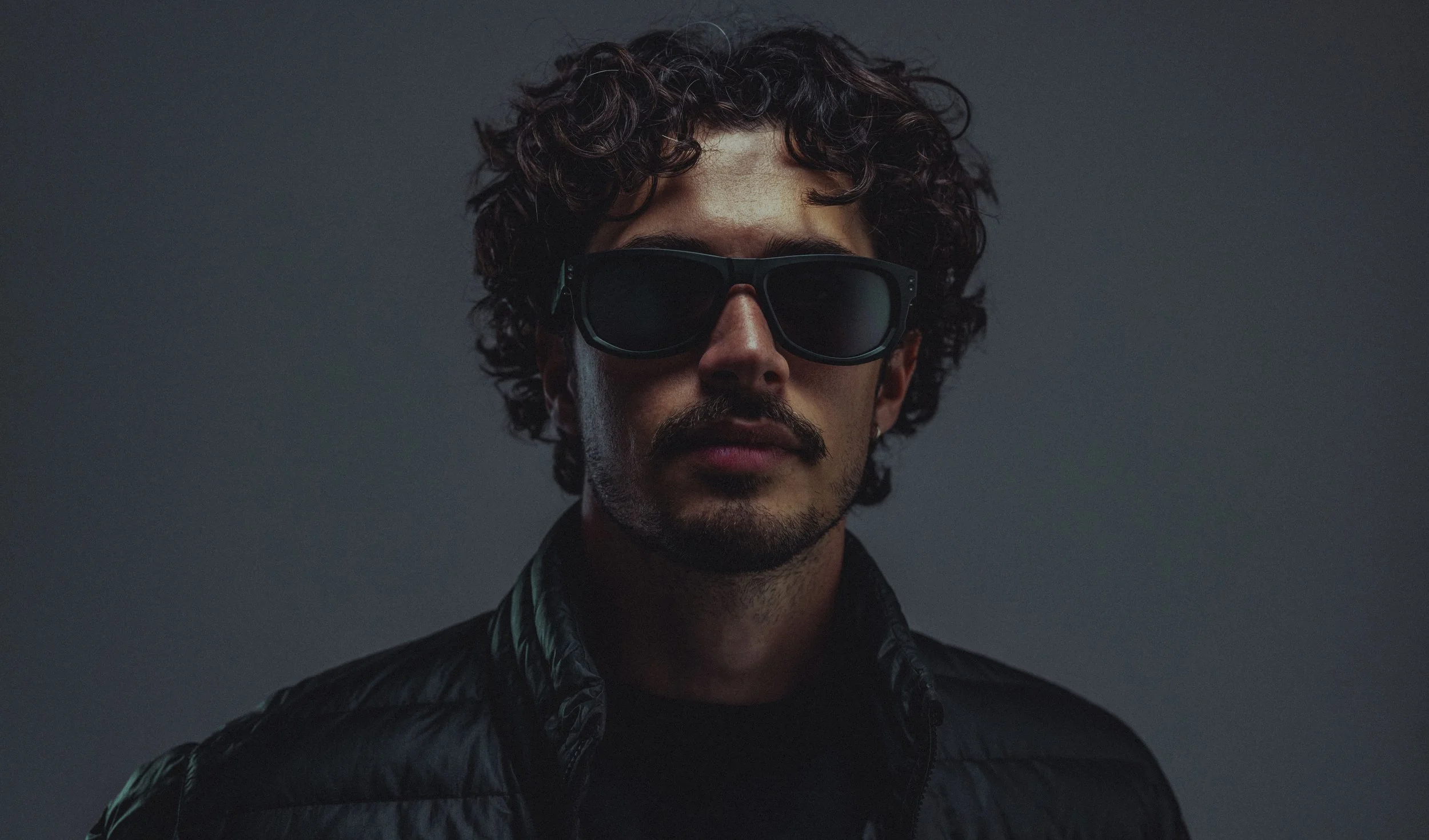 A man with curly dark hair, wearing sunglasses and a black jacket, posing against a gray background.