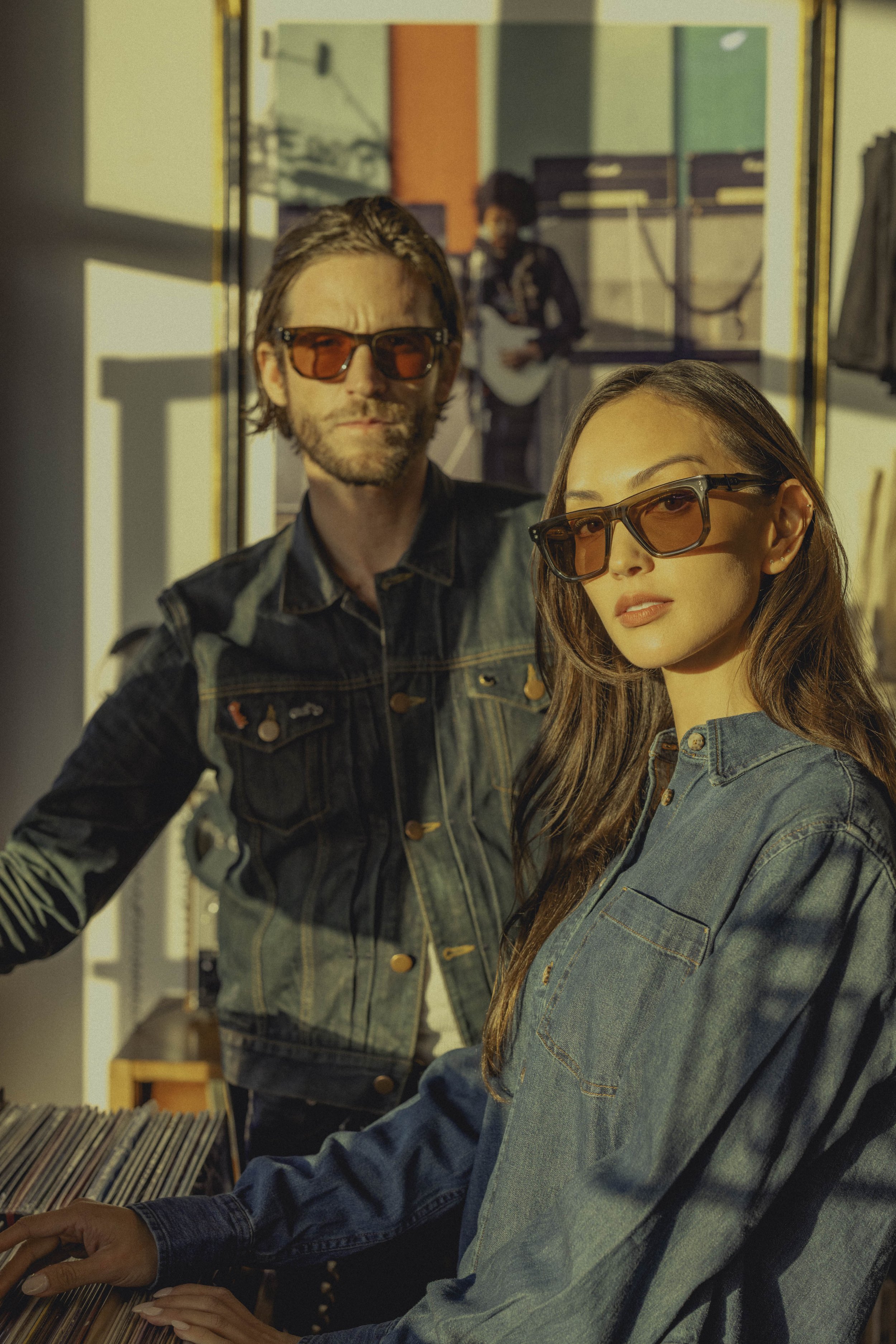 Two people, a man and a woman, wearing sunglasses and denim jackets, are taking a selfie in front of a mirror. In the background, a person is playing a guitar in a colorful room.