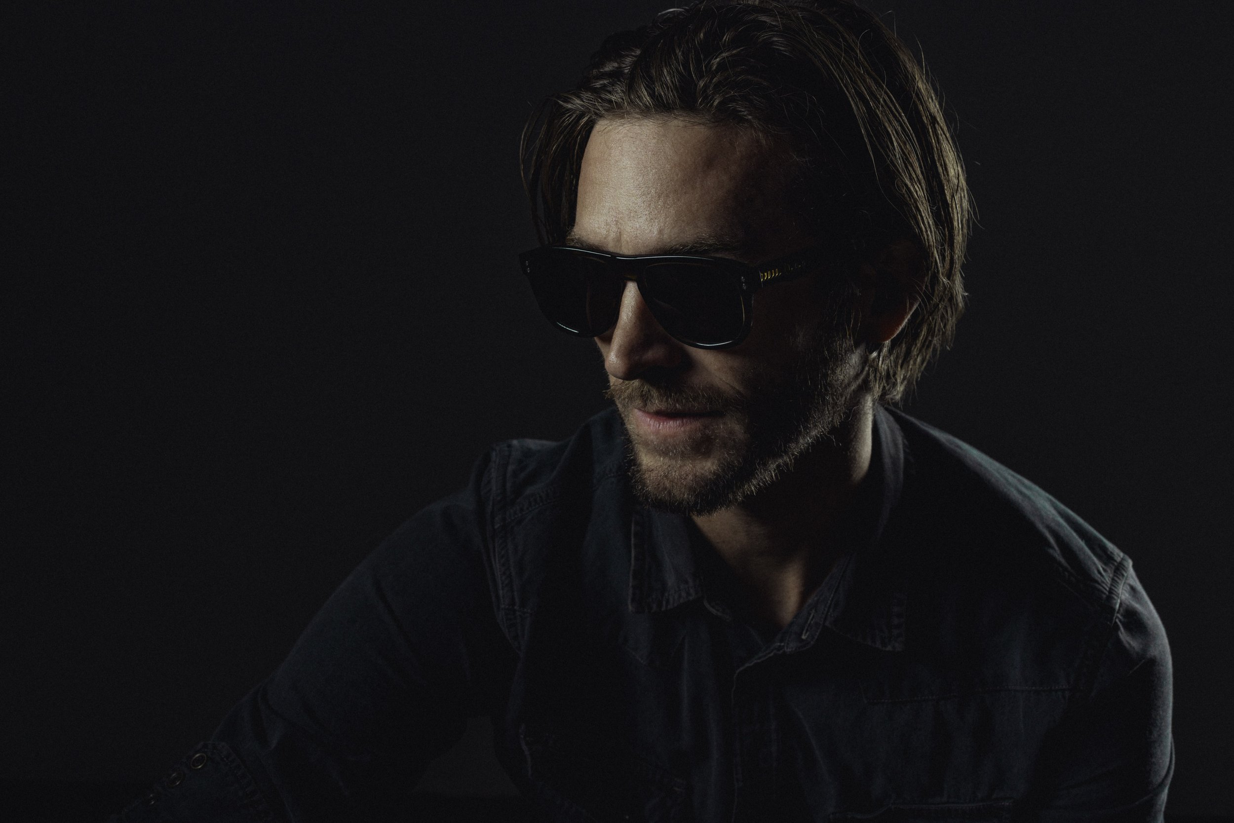 A man with brown hair and a beard wearing dark sunglasses and a dark denim jacket, looking to his left against a black background.