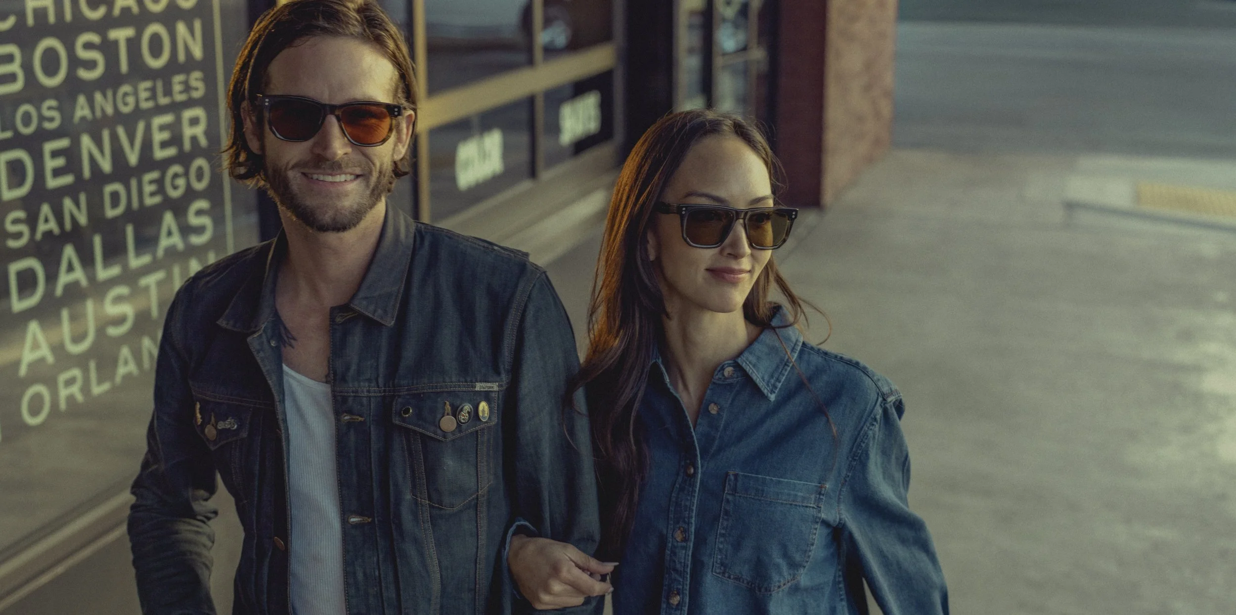 A man and a woman walking together on a sidewalk outdoors, both wearing sunglasses and denim jackets, with a city street and a sign listing various US cities in the background.