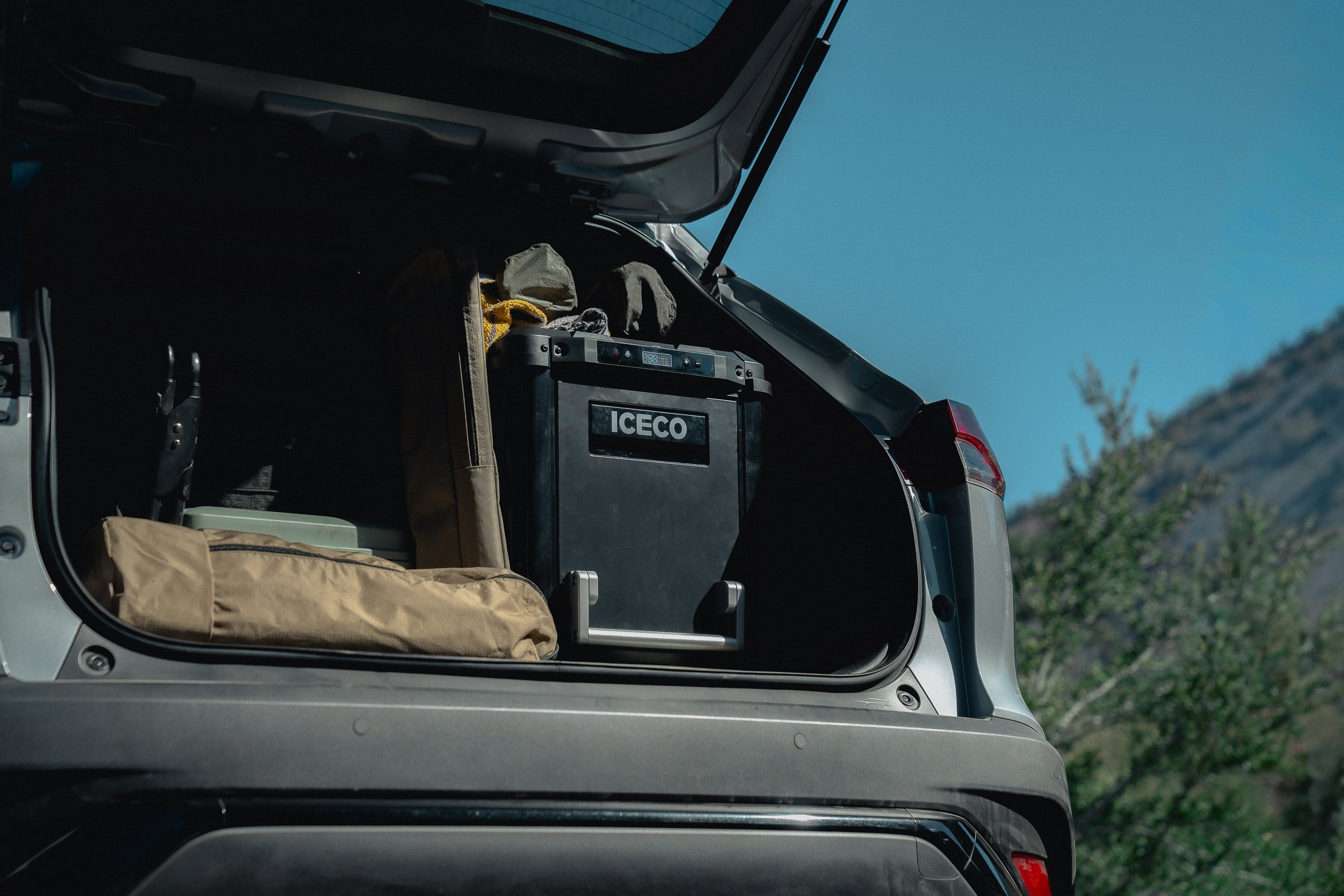 View inside the open trunk of a vehicle, showing a black ice cooler, a backpack, and some clothing items against a backdrop of blue sky and distant hillside.