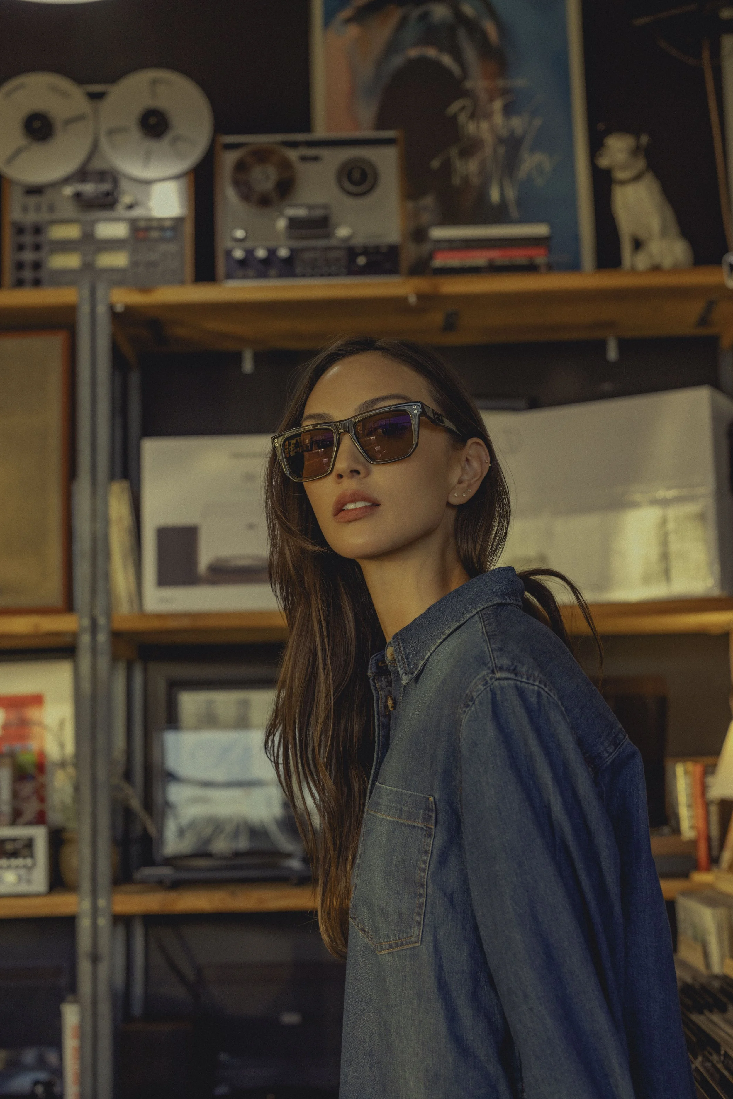 A woman with long brown hair wearing sunglasses and a denim jacket standing indoors in front of a wooden shelf filled with various vintage audio equipment and books. The background includes posters and a dog statue.
