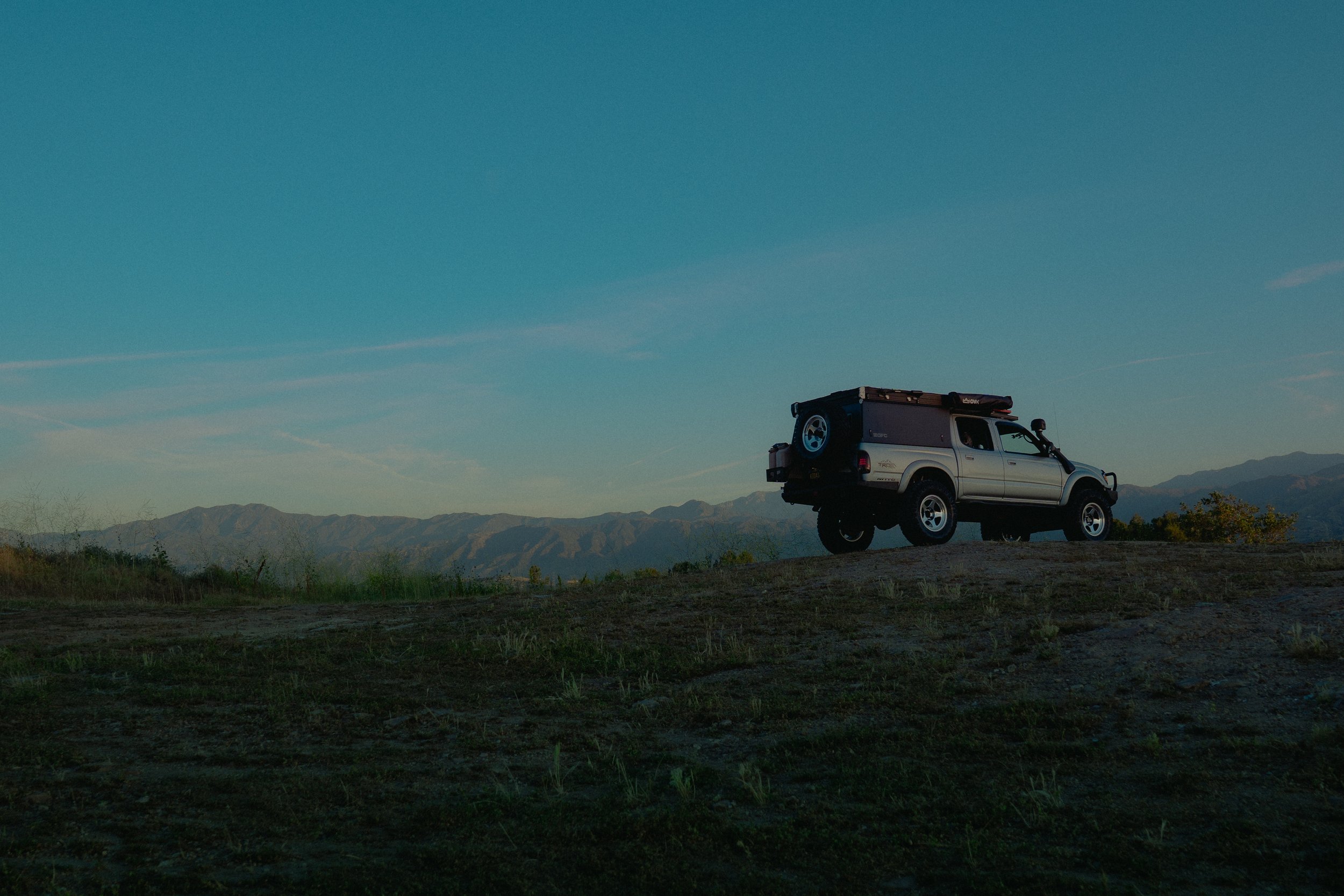 A white pickup truck with a camper shell is parked on a hilltop with mountains in the background and a clear blue sky.