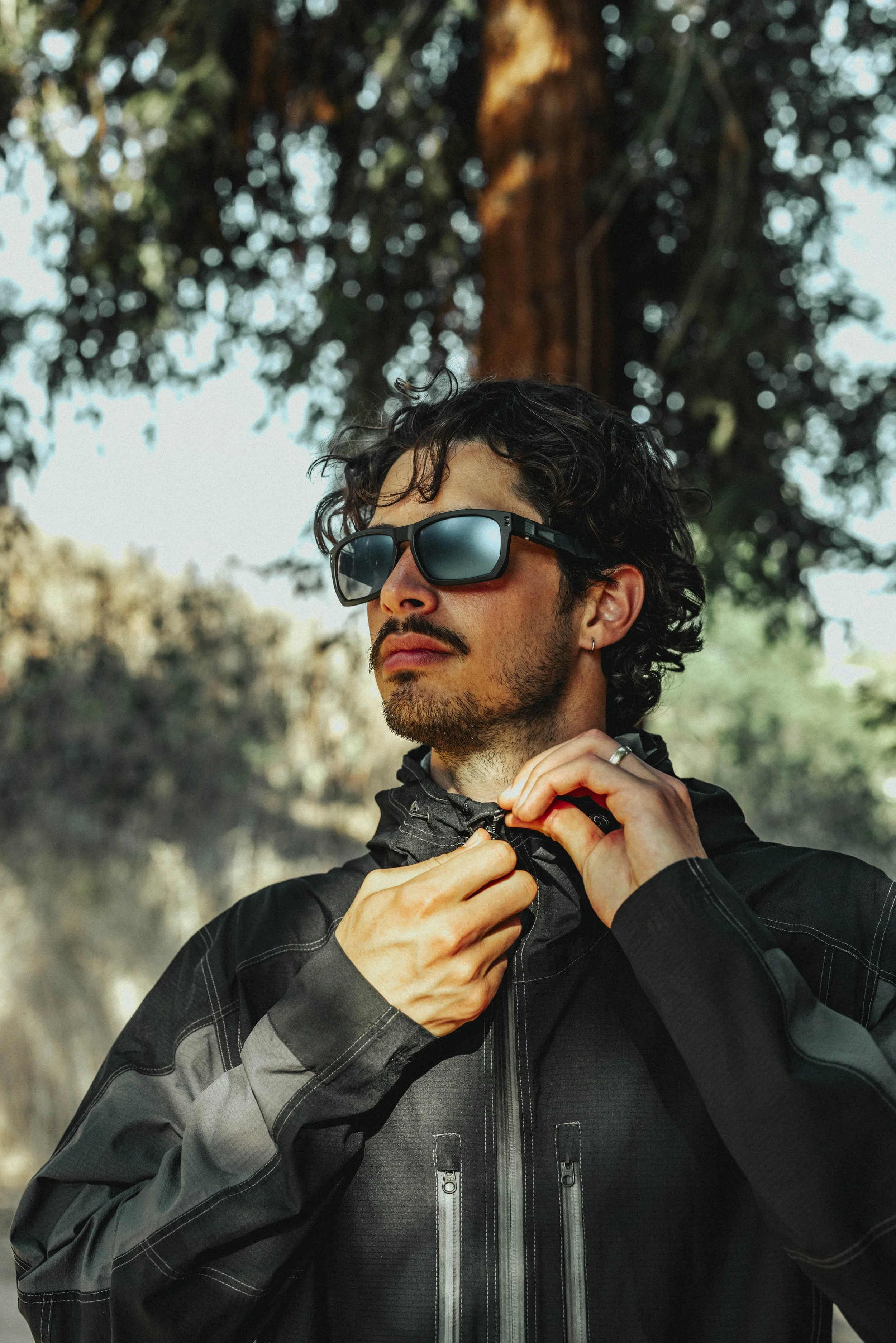 A man with curly dark hair, wearing sunglasses and a black jacket, is outdoors adjusting his jacket against a background of trees and sunlight.