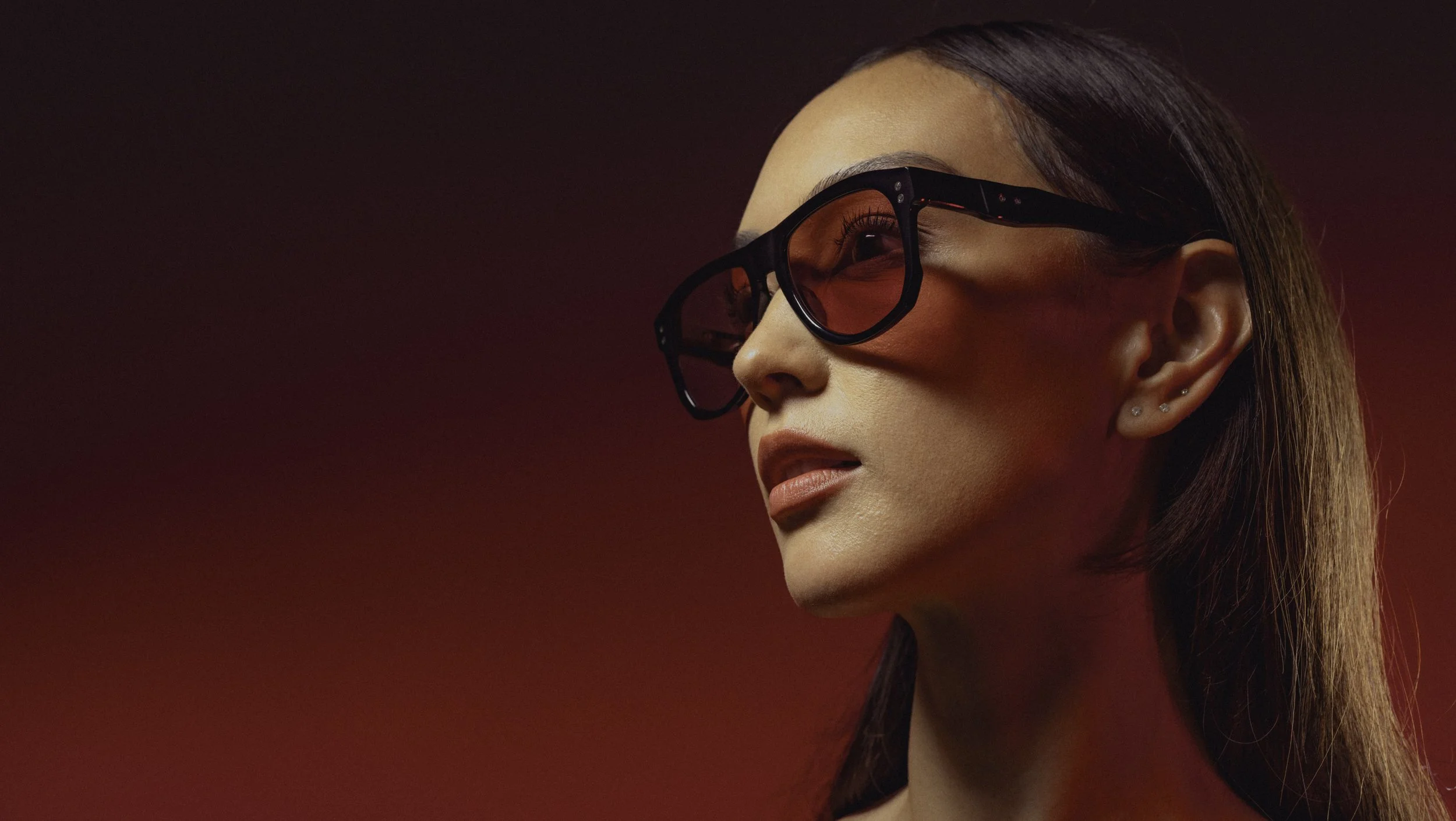 A close-up of a woman with long brown hair wearing black sunglasses with red-tinted lenses against a red background.