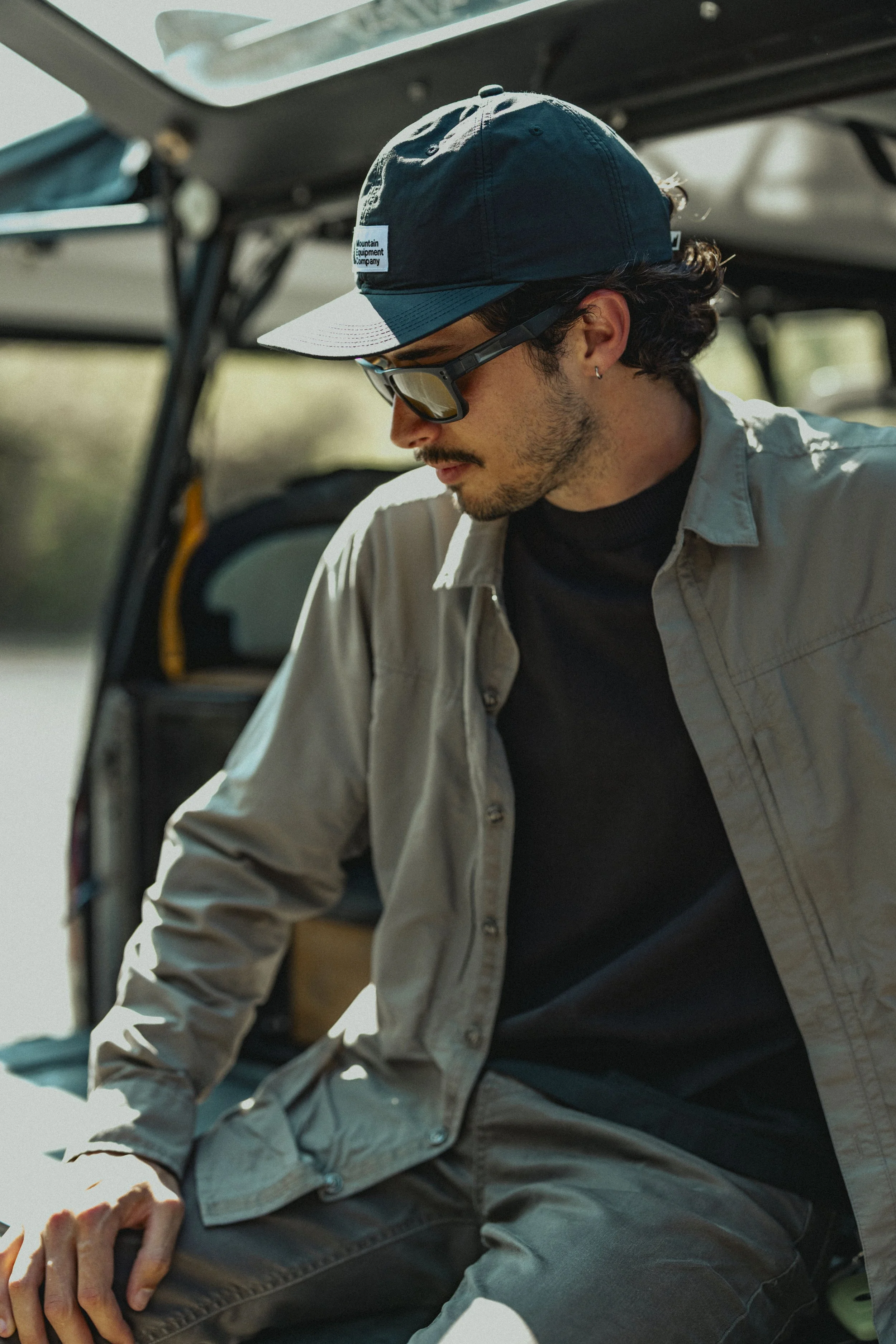 A man with dark hair, sunglasses, and a gray cap sits near a vehicle with its trunk open, wearing a gray jacket over a black shirt.