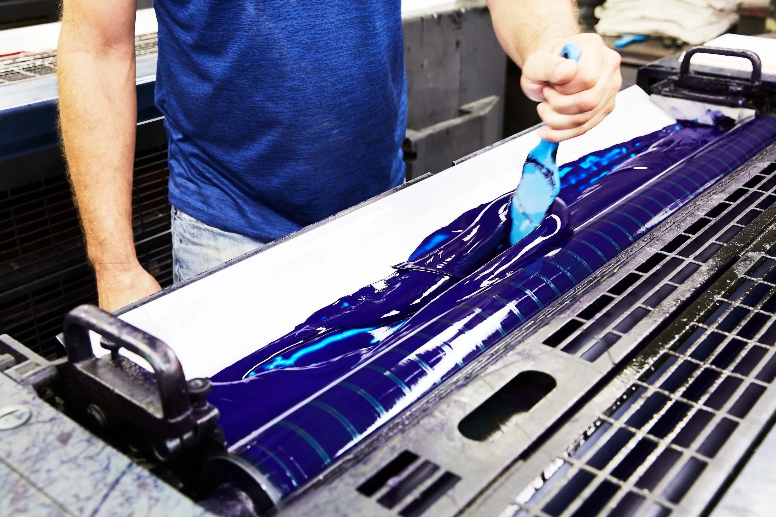 Proprint production employee applying deep blue printing ink to a press roller using a spatula, preparing the equipment for printing.
