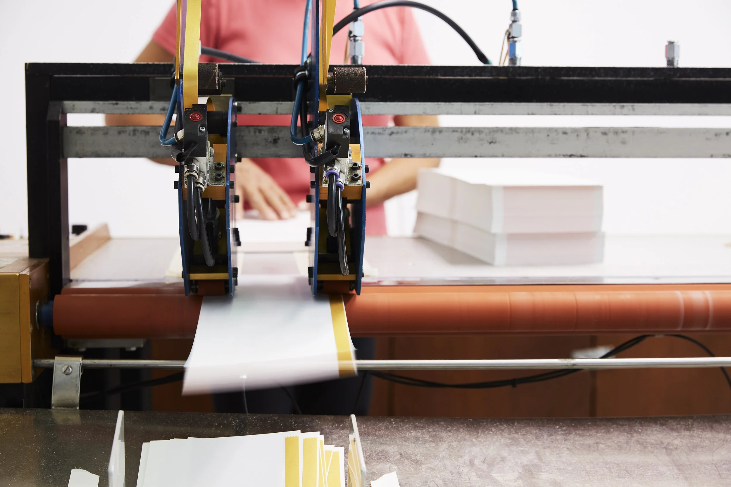 Proprint production employee working behind an industrial finishing machine as white printed material passes through rollers, with stacks of materials in the background.