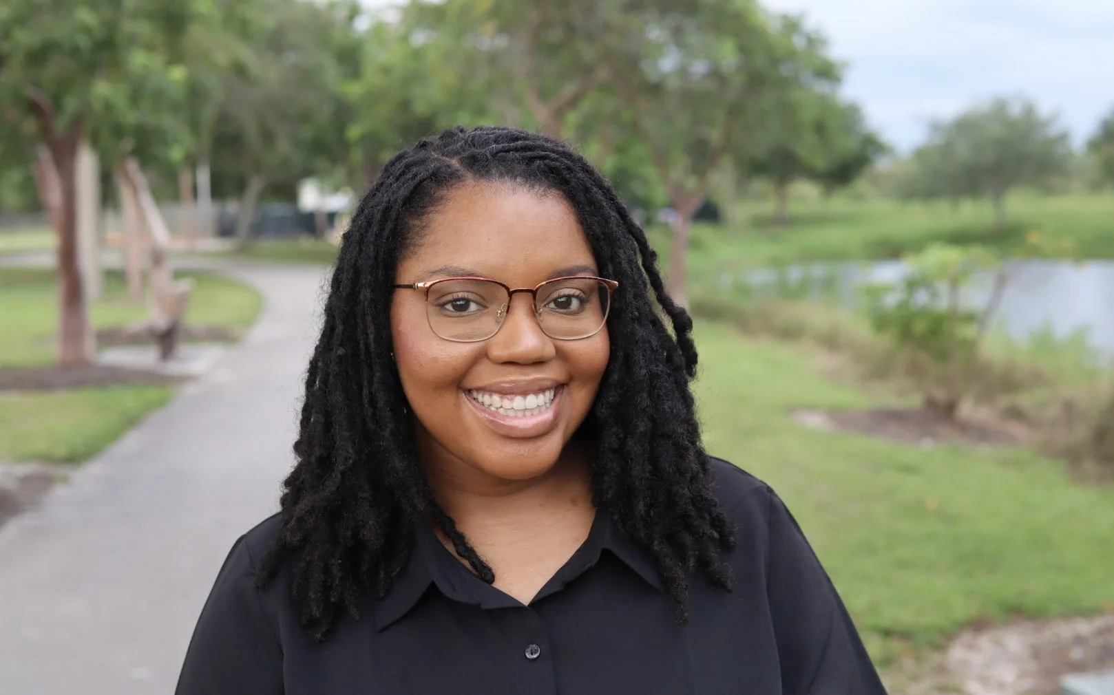 A smiling woman with glasses and dreadlocks stands outdoors in a park with trees and a pond in the background.