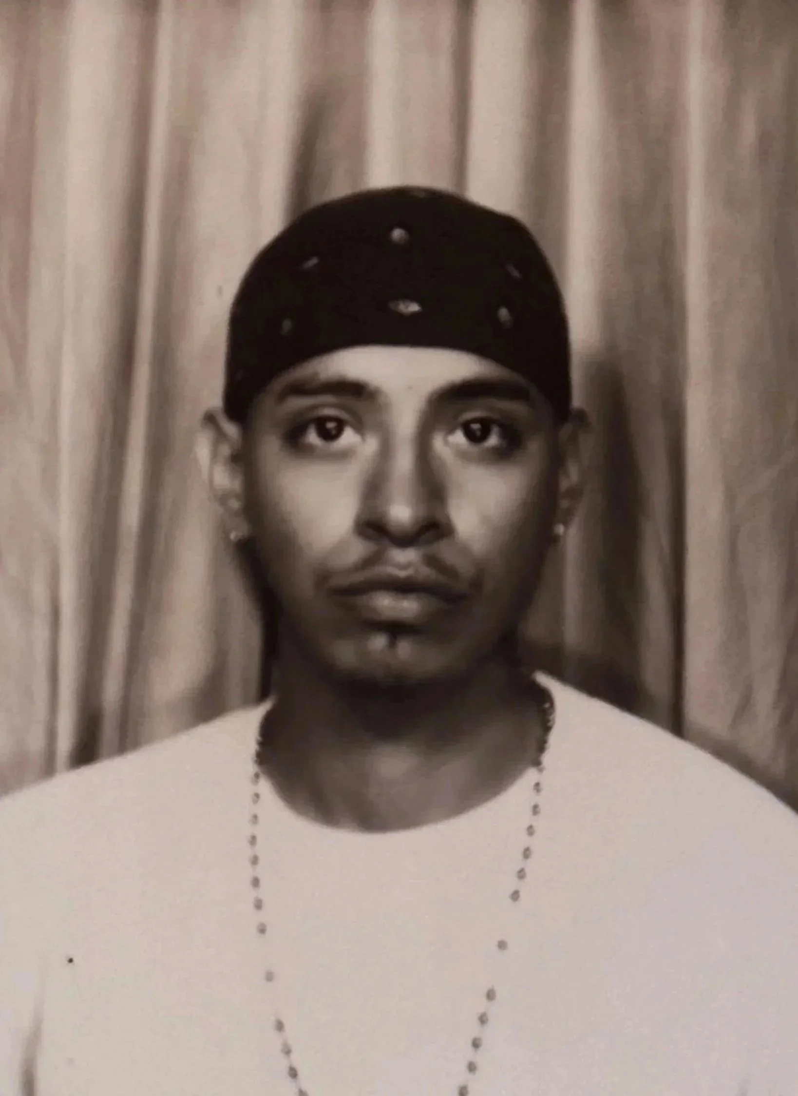 Black and white portrait of a young man wearing a bandana and a chain necklace, standing in front of a curtain.