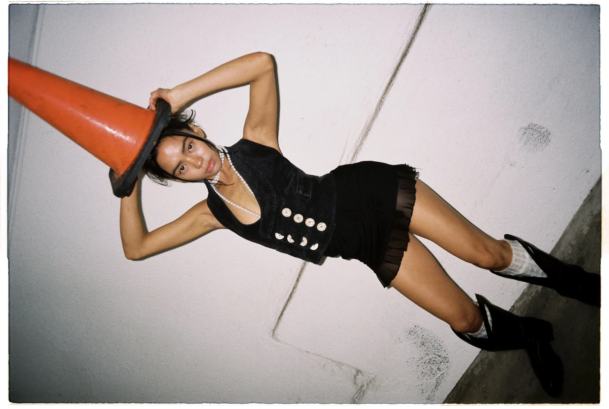 A young woman with black hair, wearing a black sleeveless vest with buttons, black shorts with sheer ruffle trim, black high-heeled boots, a pearl necklace, and a white beaded necklace, is standing against a white wall. She is holding an orange traffic cone on her head with one hand, and her other arm is bent with her hand resting on top of her head. She has a serious expression on her face.