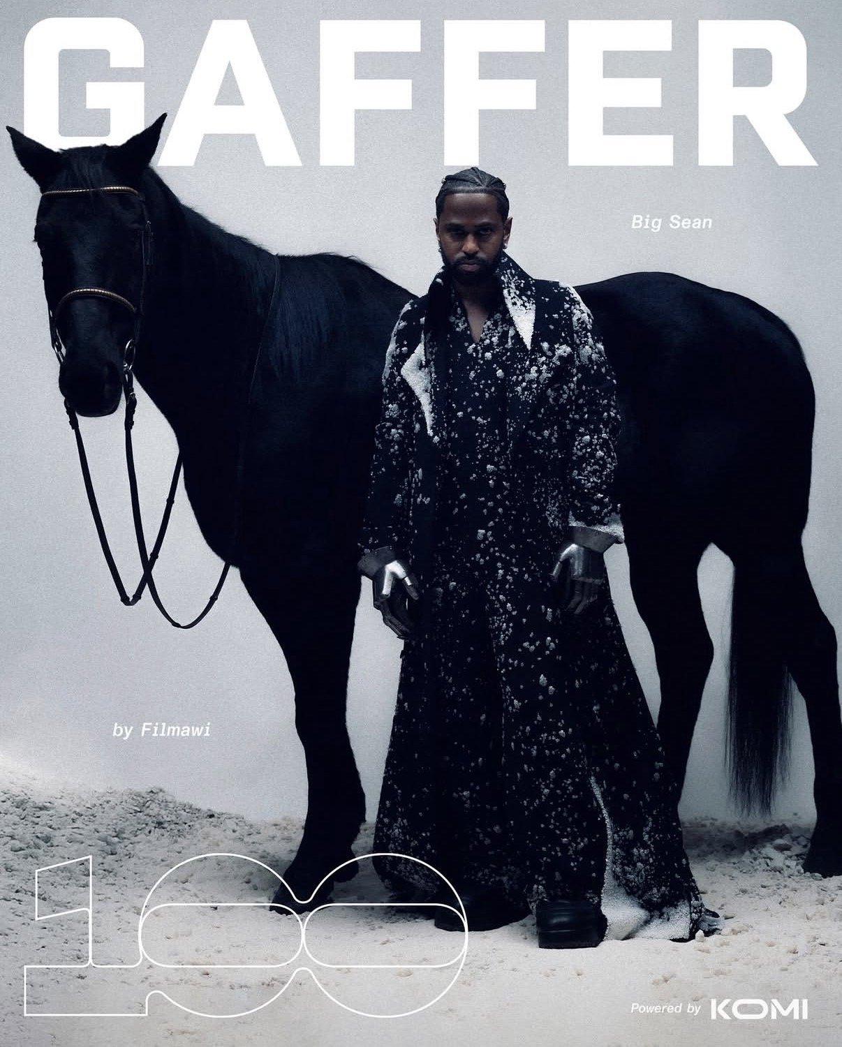A man dressed in a black and white speckled outfit standing next to a black horse against a plain white background.