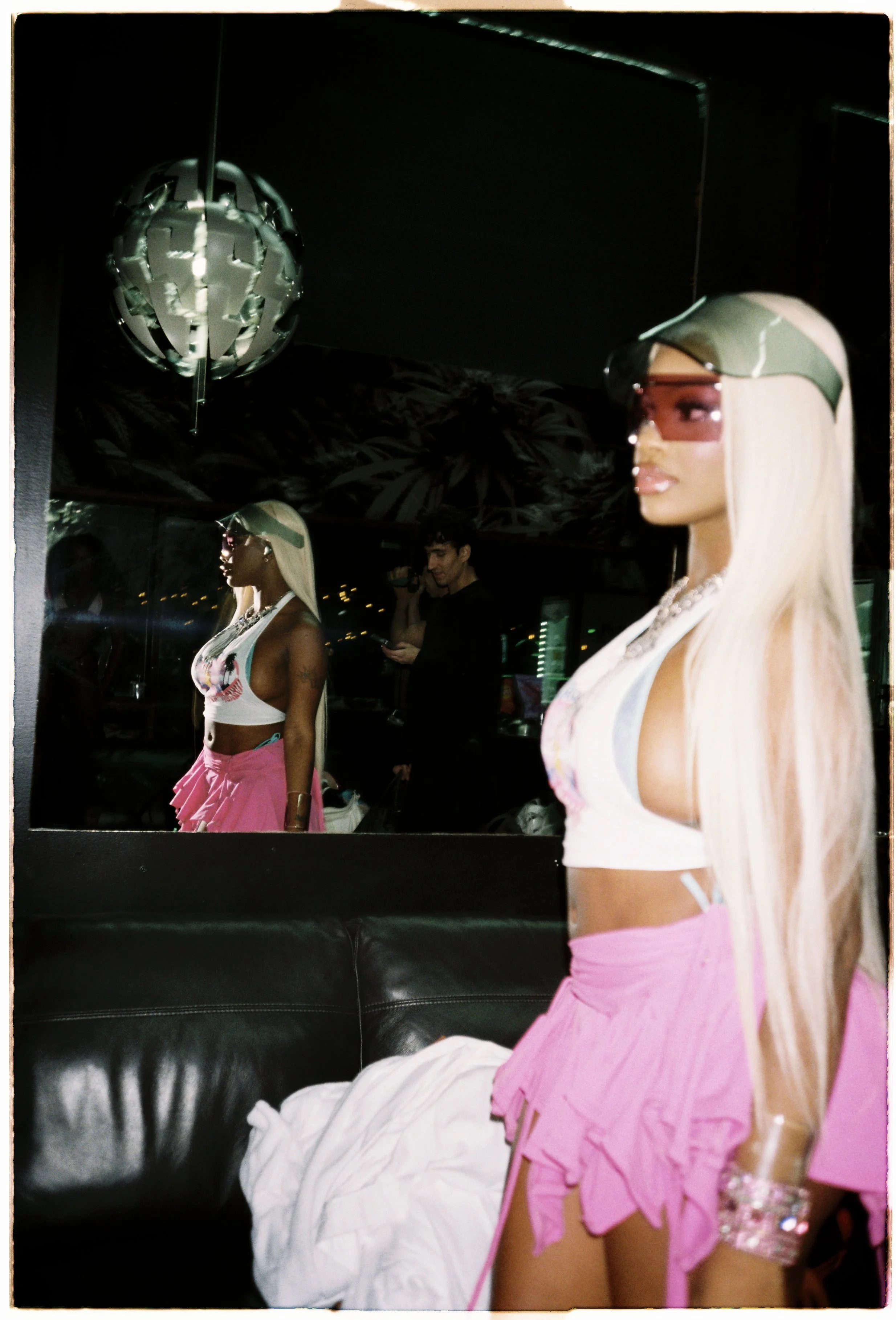 A woman with long blonde hair, wearing sunglasses, a white crop top, and a pink skirt, sitting in a dimly lit room with a mirror reflecting the mannequin and a man taking a photo.