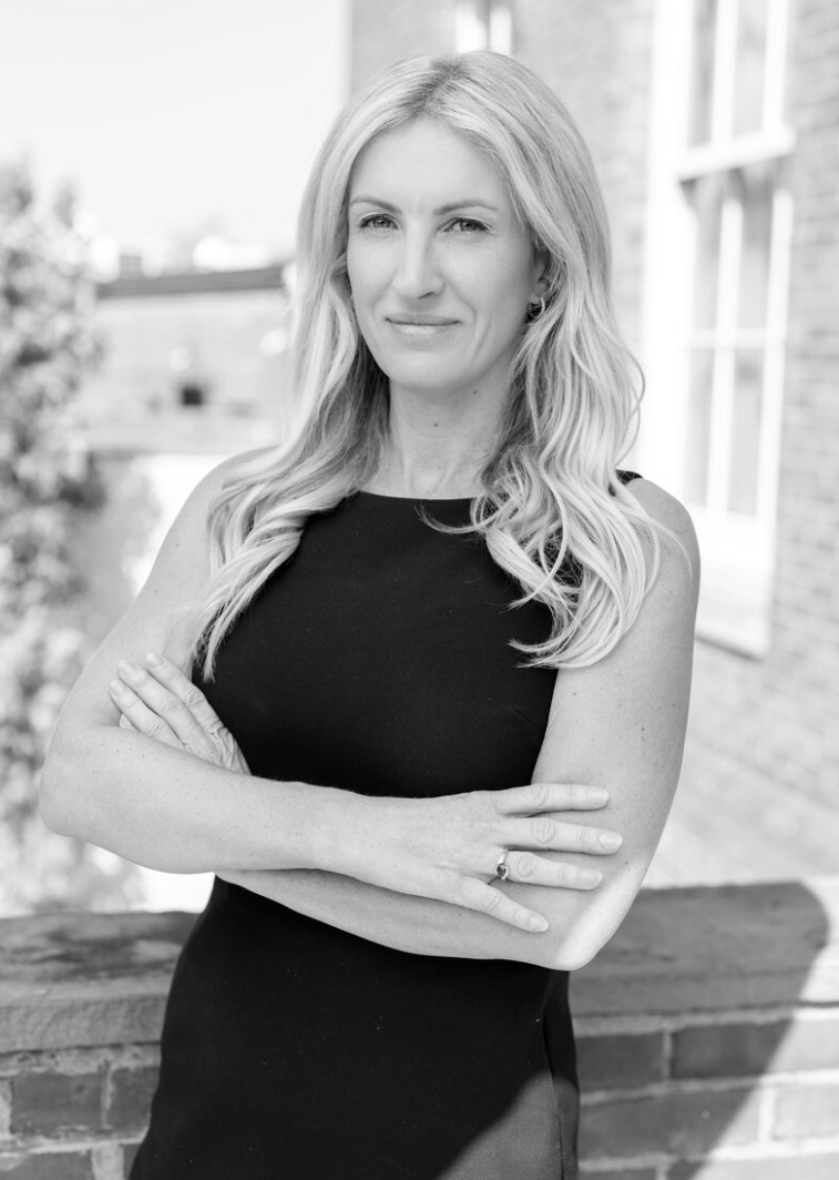 A black-and-white photo of a woman with long, wavy blonde hair, wearing a sleeveless black dress, standing outdoors with her arms crossed, in front of a brick building and window.