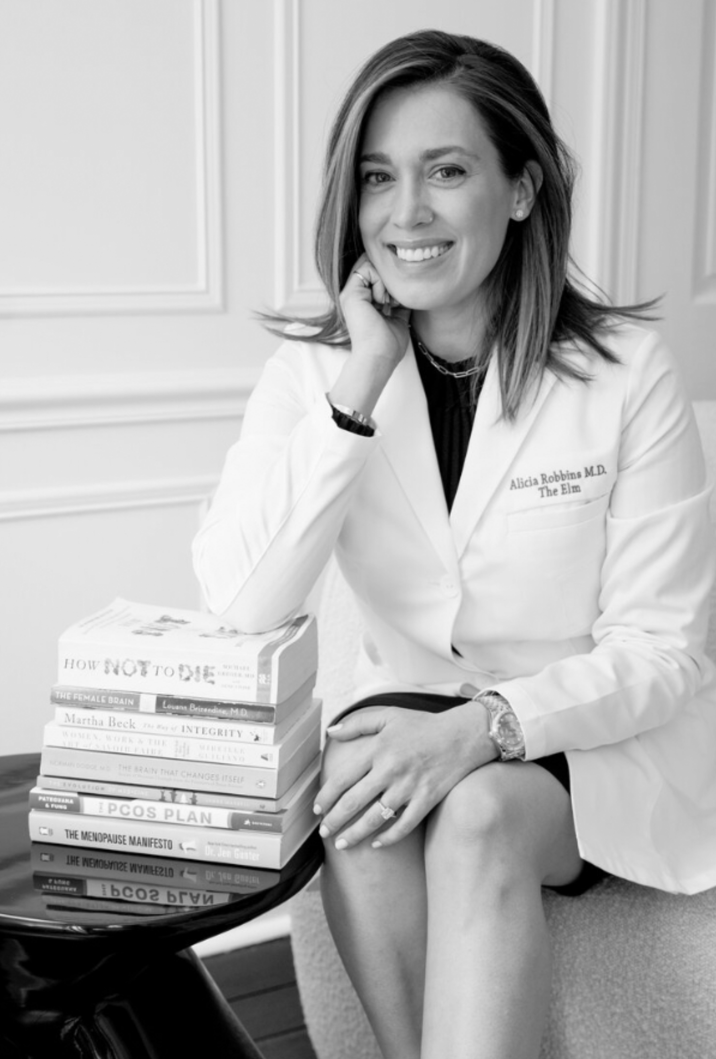 A woman in a white lab coat sitting indoors with a stack of books on a small table next to her, smiling at the camera.
