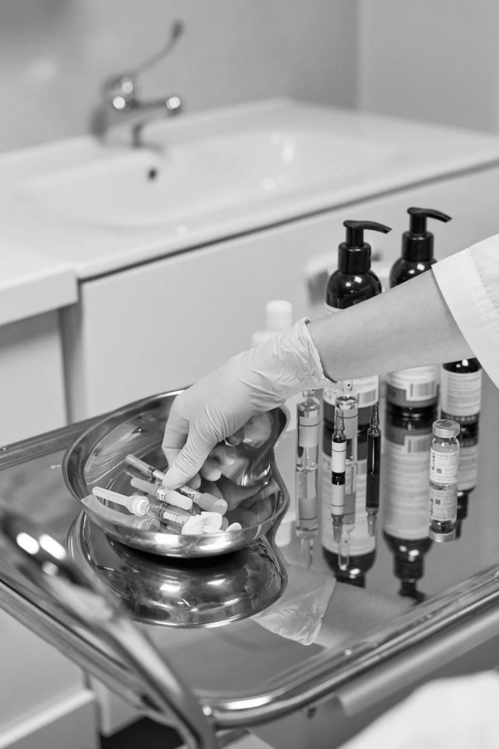 A healthcare worker's gloved hand placing syringes into a metal tray, with various medicine bottles nearby in a clinical setting.