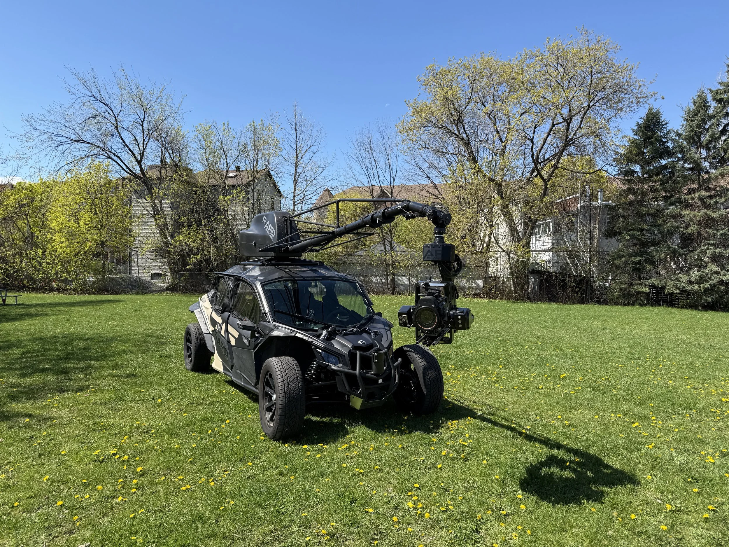 A small futuristic vehicle with a large camera mounted on top, parked on a grassy field with trees and houses in the background under a bright blue sky.