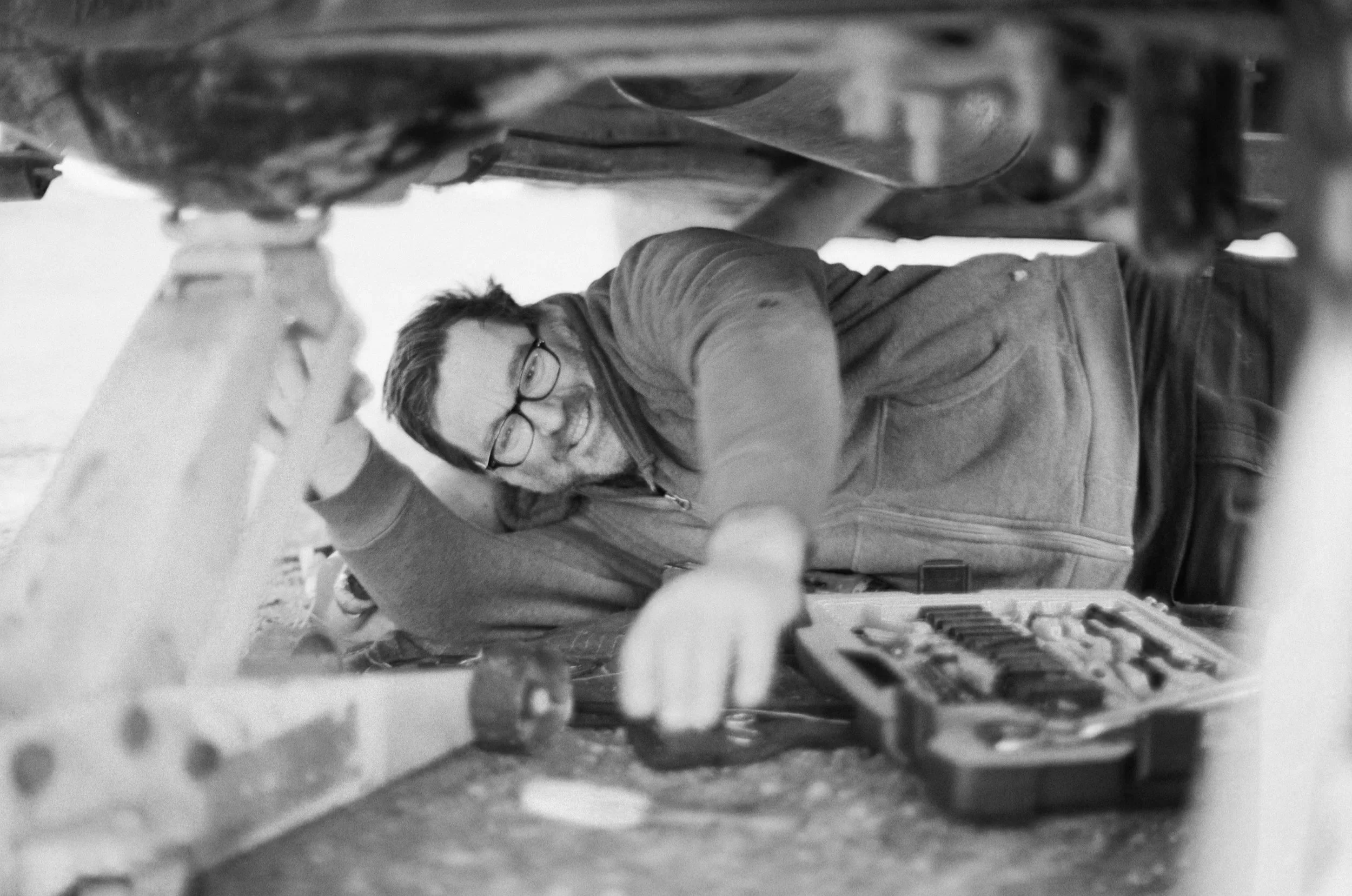 Man working under a vehicle, lying on the ground, using a red toolbox with various tools, smiling, and wearing glasses and a gray hoodie.