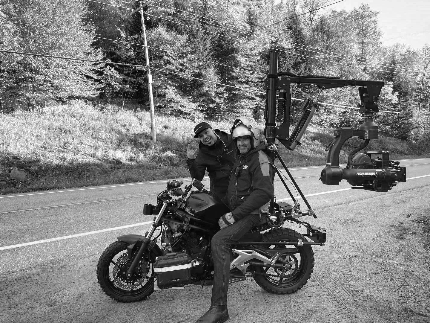 Two men with motorcycle-mounted camera equipment on a rural roadside, one wearing a helmet and the other making a peace sign, with fall foliage background.