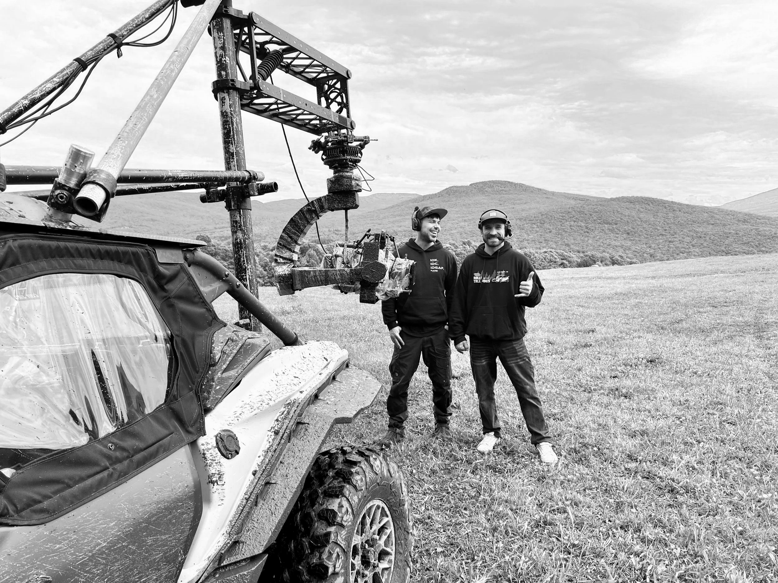 Two men standing in a grassy field with green hills and mountains in the background, posing beside a vehicle equipped with filming equipment, one giving a thumbs-up.