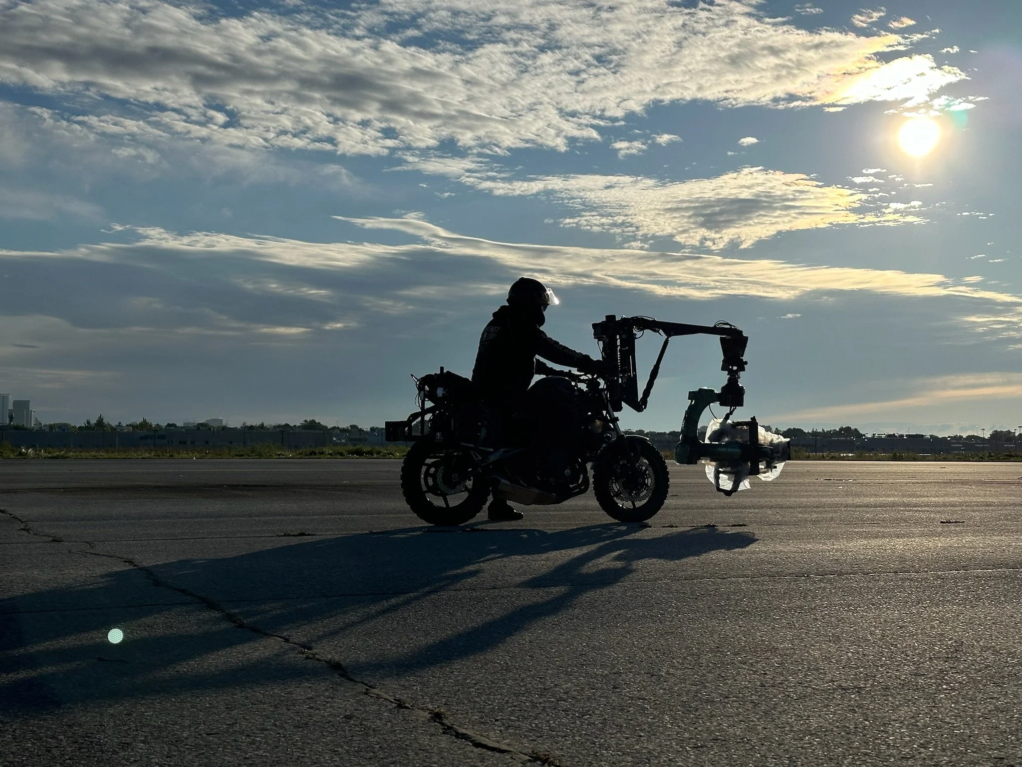 Silhouette of a person riding a motorcycle with a mounted robotic arm on a wide open road during sunset, with a partly cloudy sky and the sun in the sky.