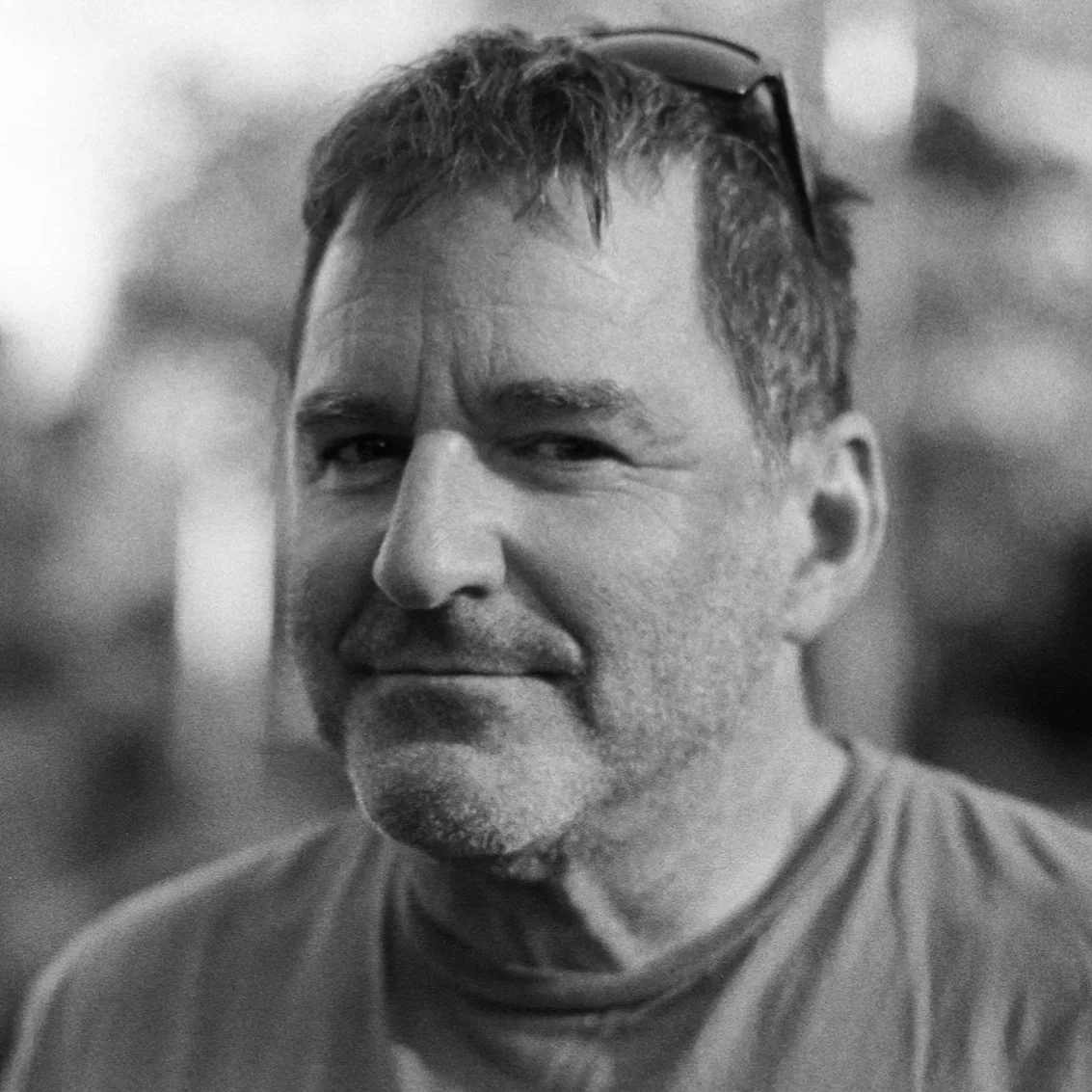 A man wearing dark sunglasses and a t-shirt, sitting in a vehicle, is smiling slightly. The reflection in his sunglasses shows a person taking a photo and part of the vehicle's interior.