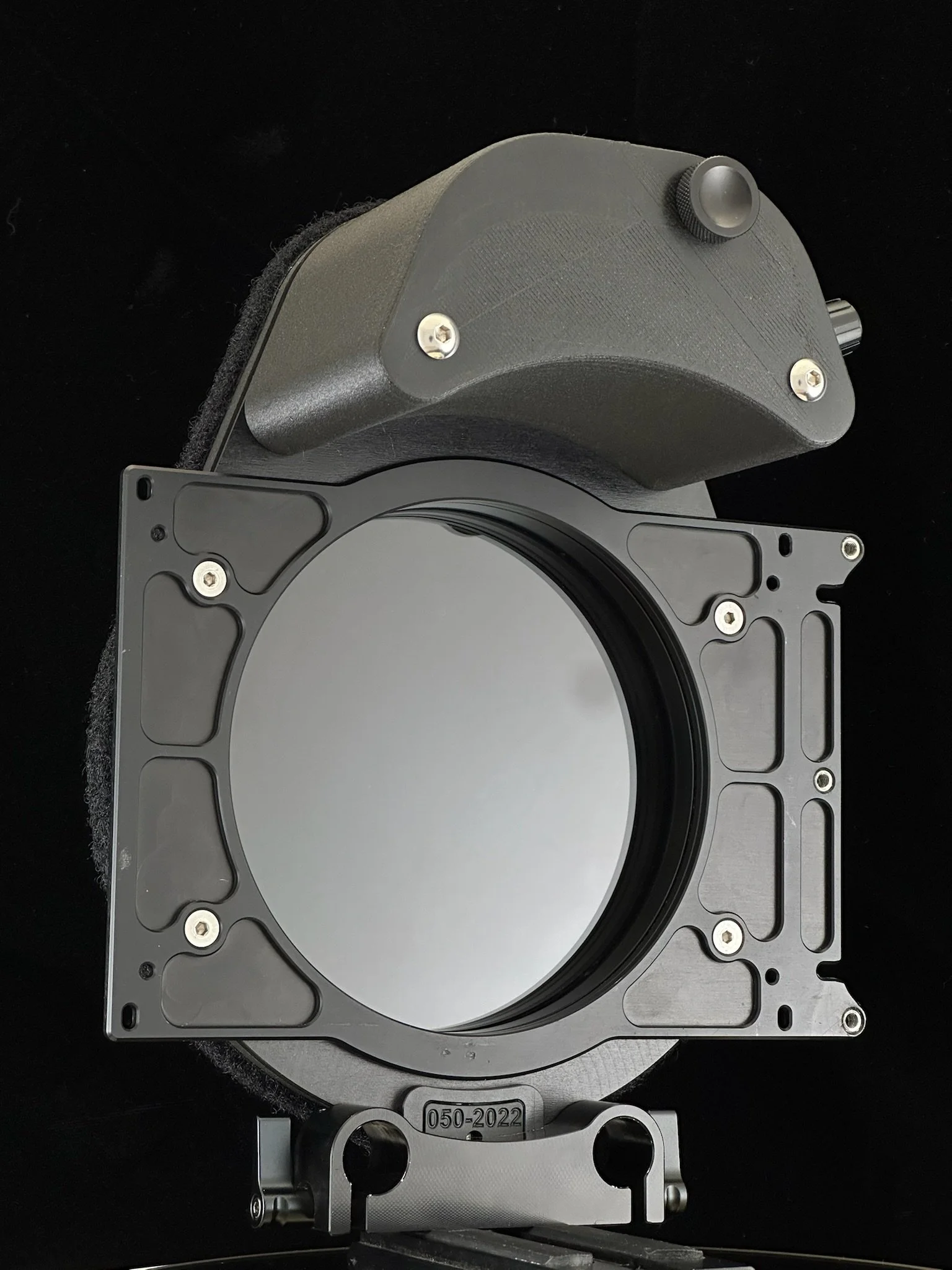 A large piece of industrial equipment, possibly a component of a scientific instrument, with a circular reflective surface and various metallic and plastic parts, set against a black background.