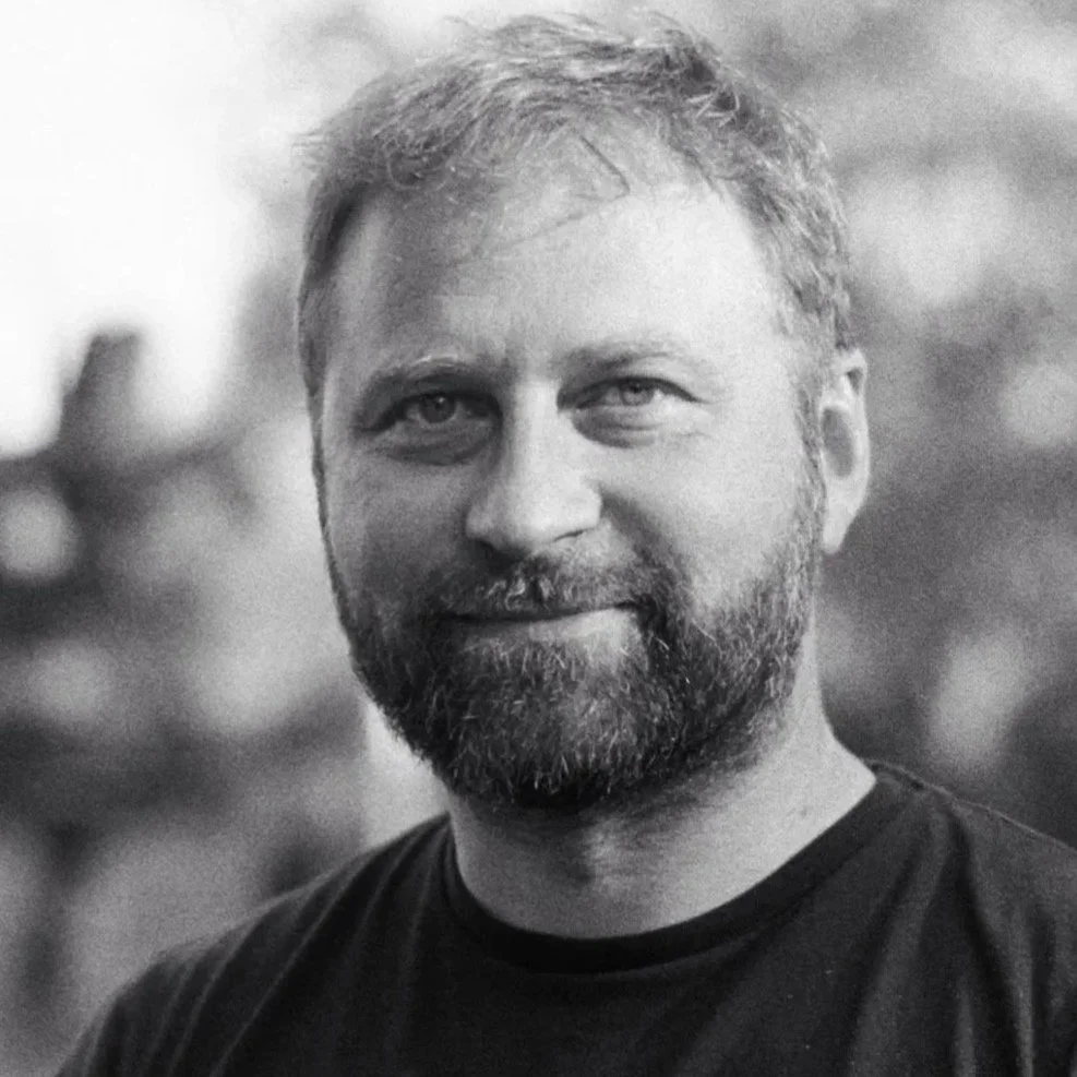 DENYS SVOIAK smiling man wearing sunglasses and a T-shirt with a car line drawing, sitting in an airport or similar setting.