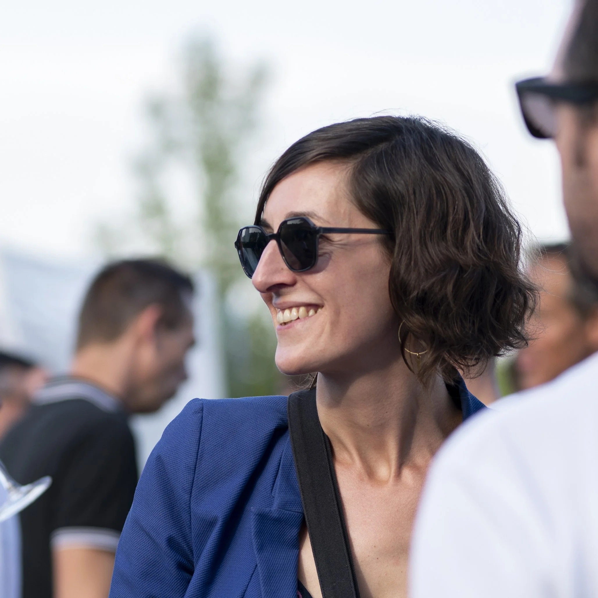 Femme souriante portant des lunettes de soleil noires, vêtue d'une veste bleue, lors d'un événement extérieur, entourée de plusieurs personnes.