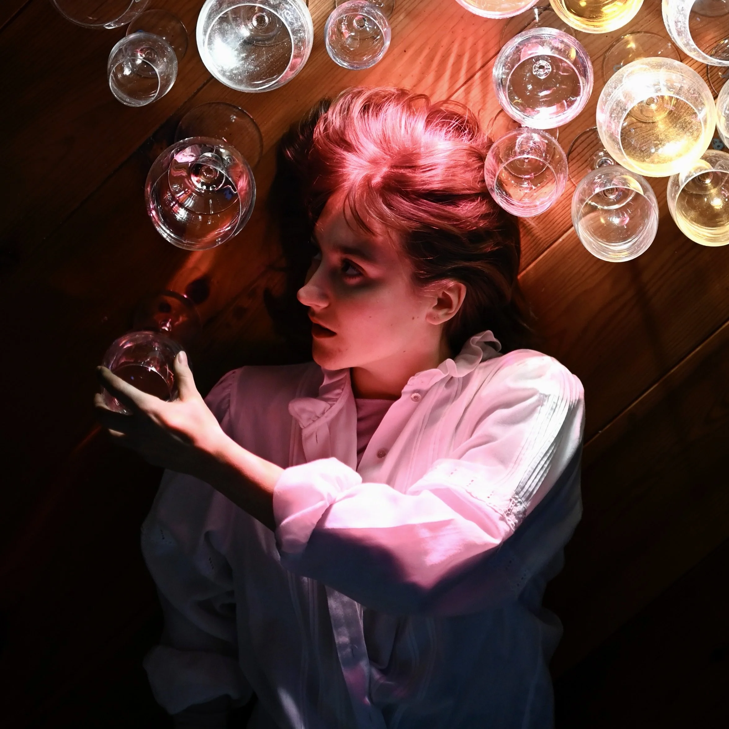 A woman with dark hair lying on a wooden floor surrounded by wine glasses filled with red and white wine, illuminated by pink and yellow lighting.