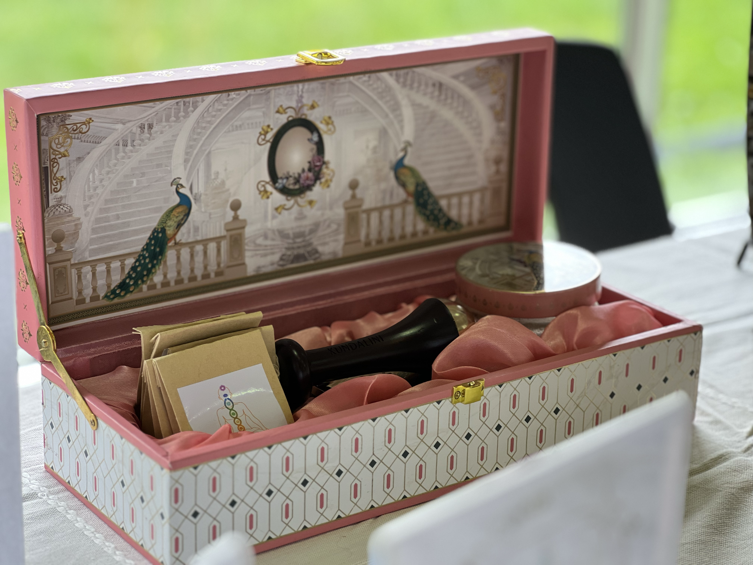 Open pink gift box with peacock design, containing a black stamp, a small stack of paper, and a round makeup container, lined with pink satin fabric, sitting on a table with a grassy background.