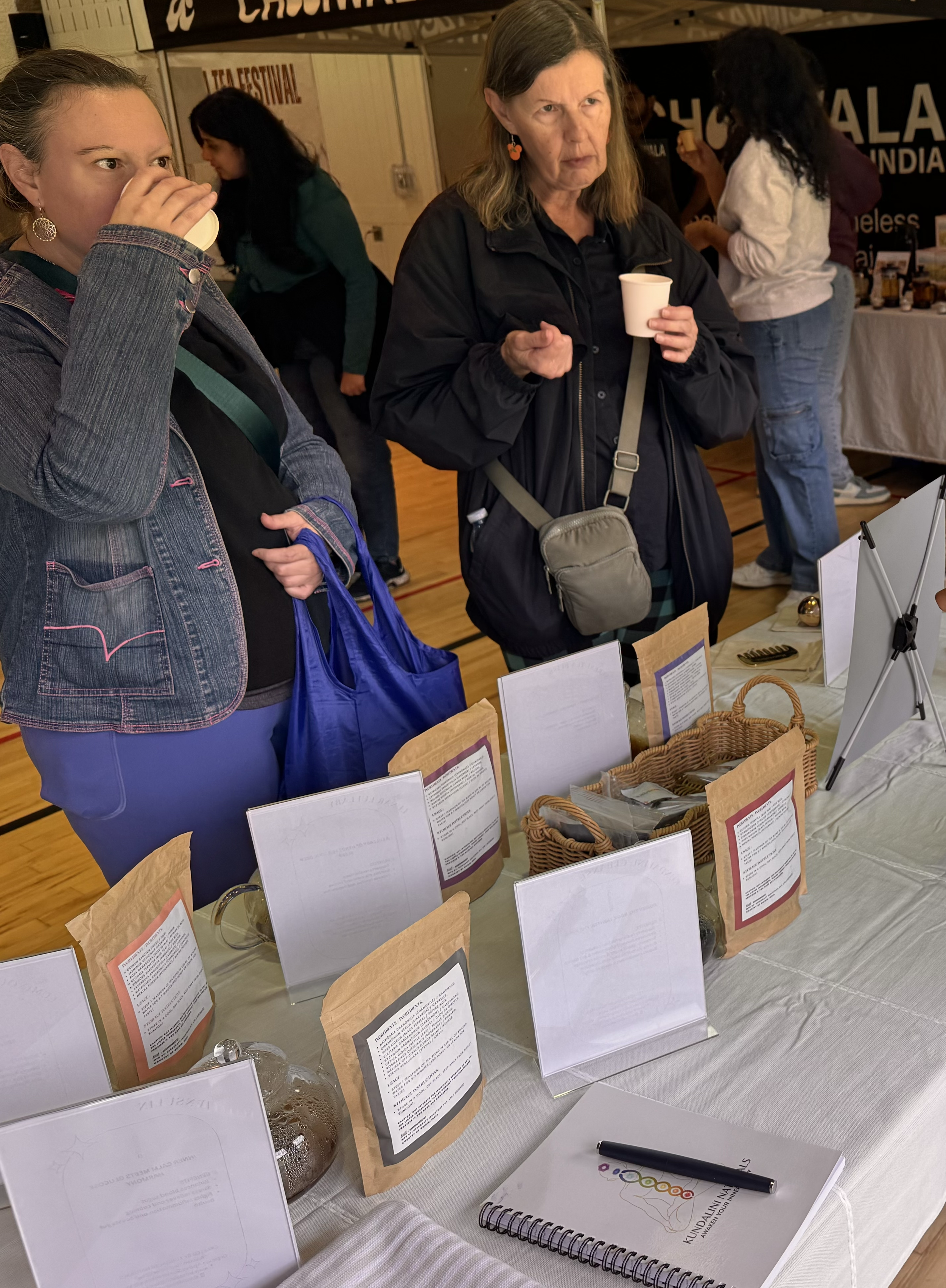 Two women are standing at a table with various products in paper and clear plastic bags, and informational cards. One woman is drinking from a paper cup, while the other is observing. There are other people in the background at what appears to be an 