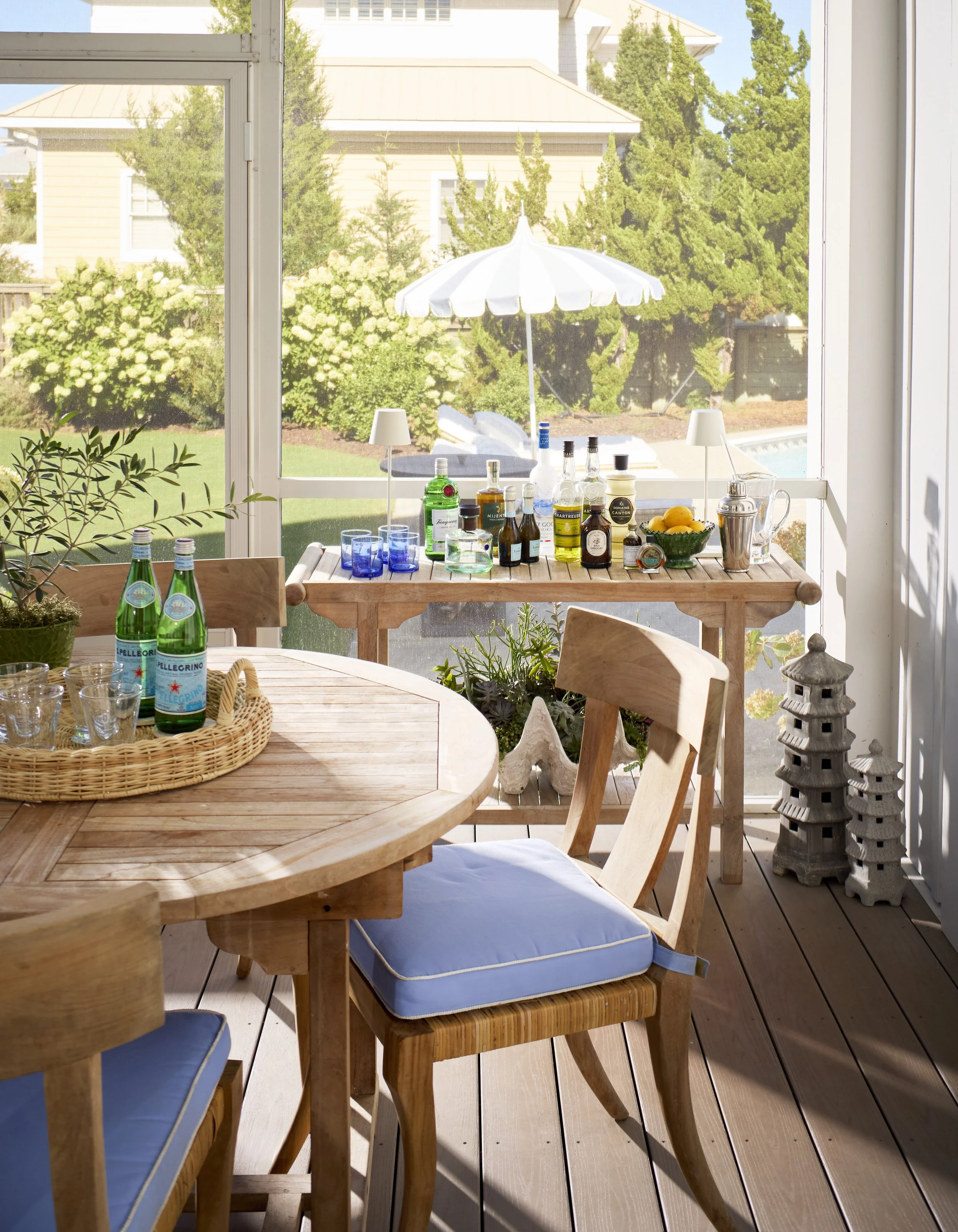 Sunlit enclosed porch with a round wooden dining table with a blue-cushioned chair, a basket with glassware, and a wooden side table with bottles, glasses, a bowl of lemons, and barware. Decor includes plants, lanterns, and an umbrella outside, overlooking a backyard with trees and a swimming pool.