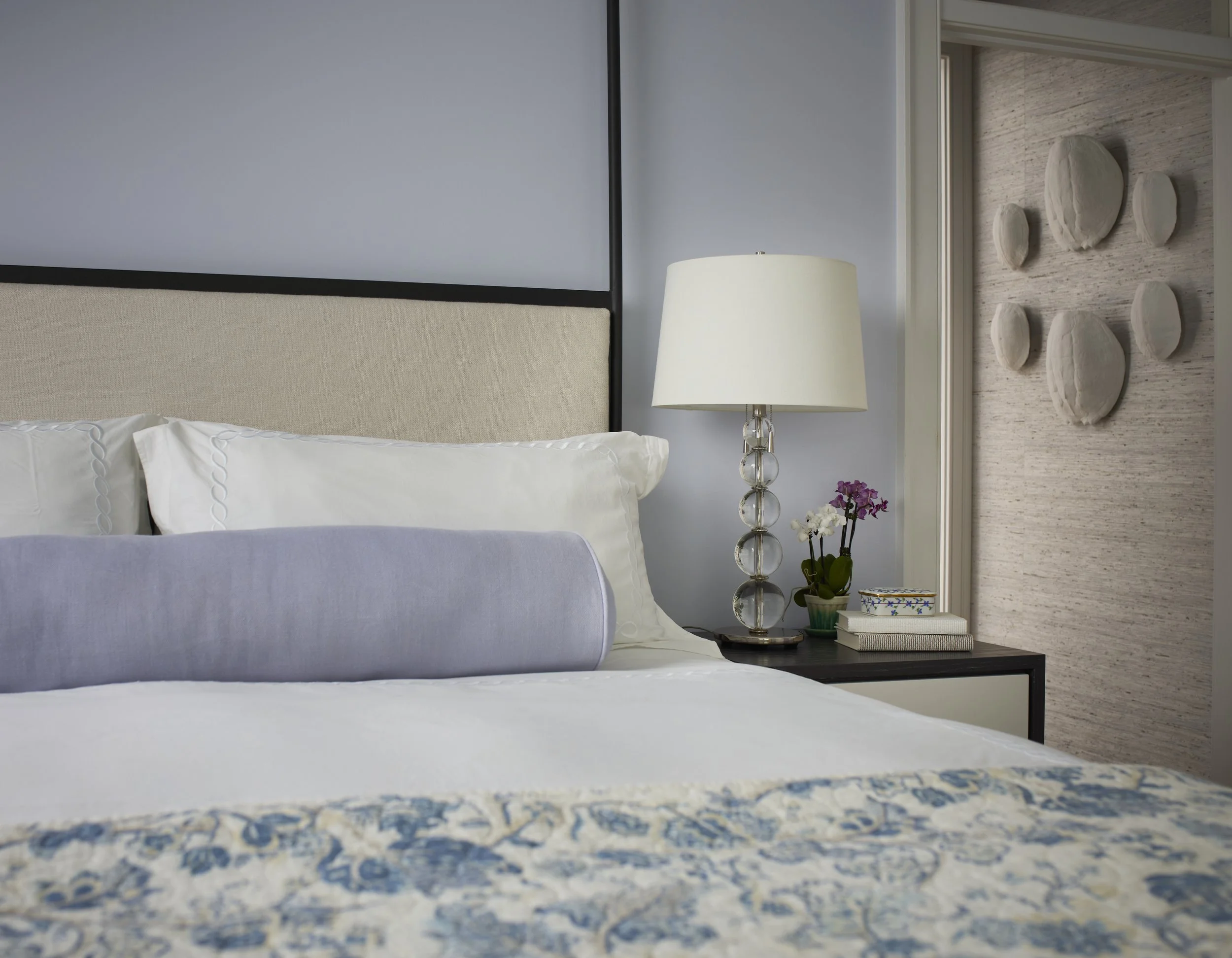 A neatly made bed with white linens and a decorative purple bolster pillow, beside a nightstand with a glass ball table lamp, a potted orchid, and a closed book, in a modern bedroom with textured wall art.