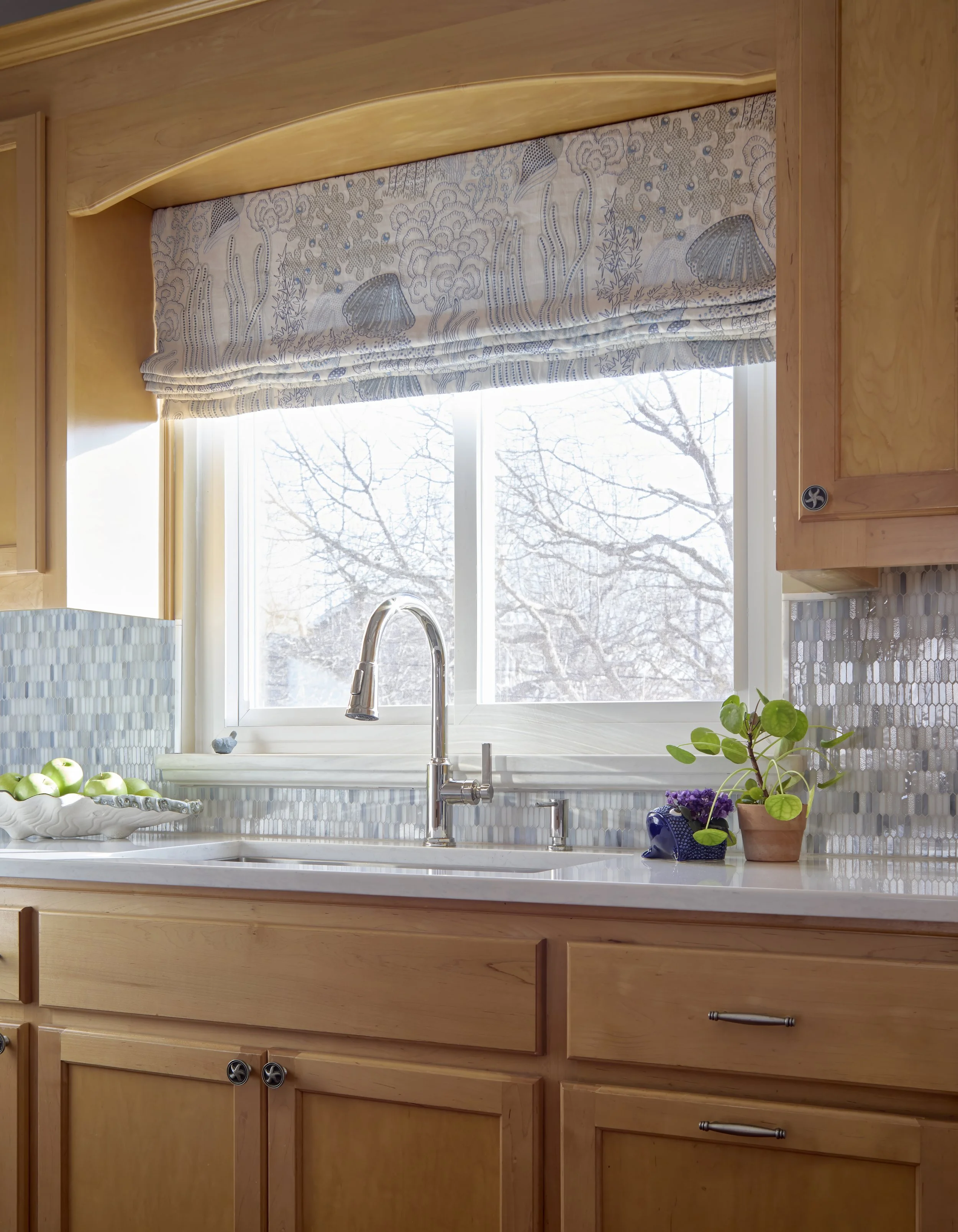 A kitchen countertop with a window above the sink, which has a silver faucet. There are green apples in a white dish on the left, and a small potted plant and a decorative ceramic object on the right. The window has a patterned valance and shows a view of leafless trees outside.