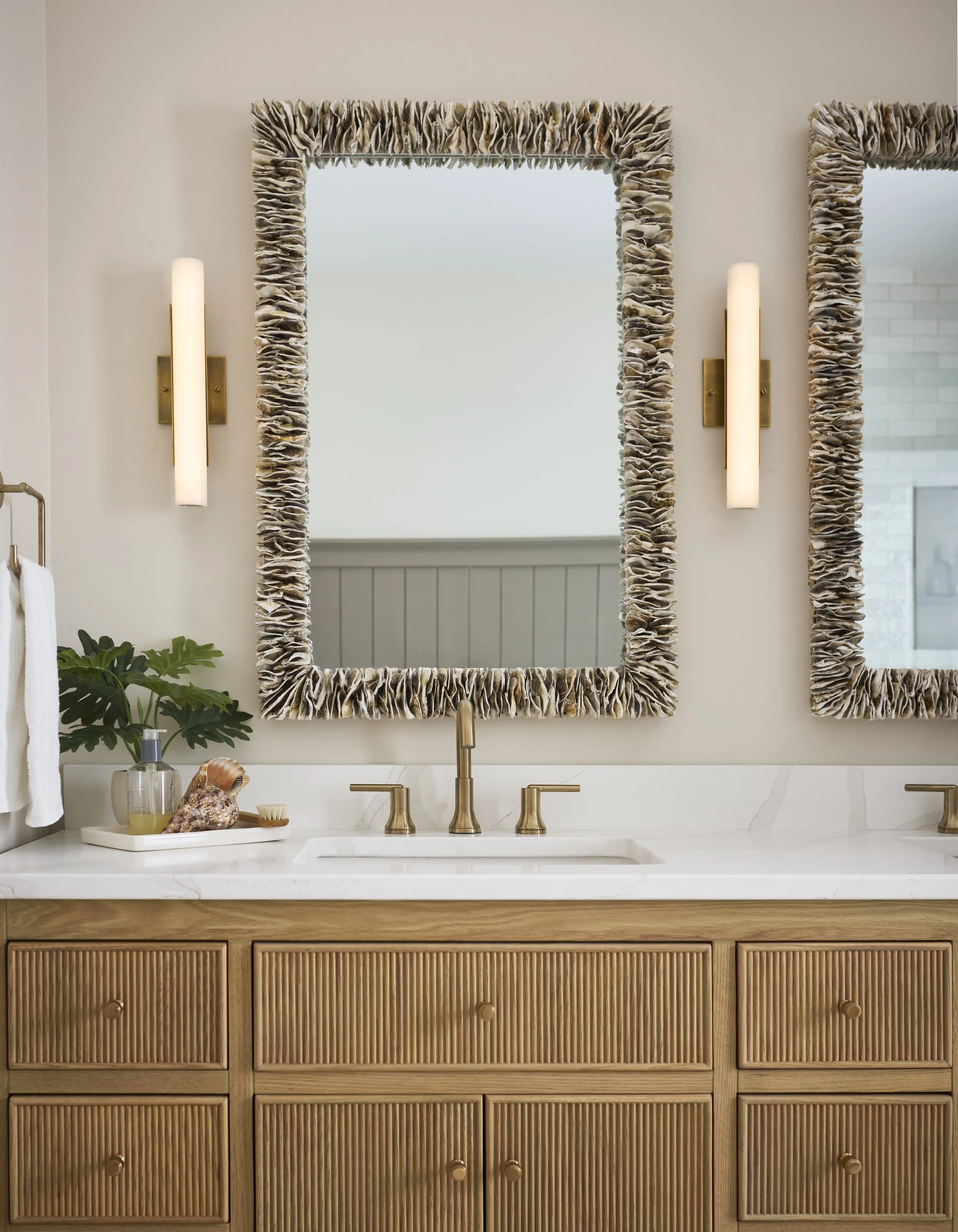 Bathroom vanity with two oyster shell mirrors, brass fixtures, marble countertop, fluted panel vanity, and decor items including a plant, soap dispenser, and decorative shell accessories.