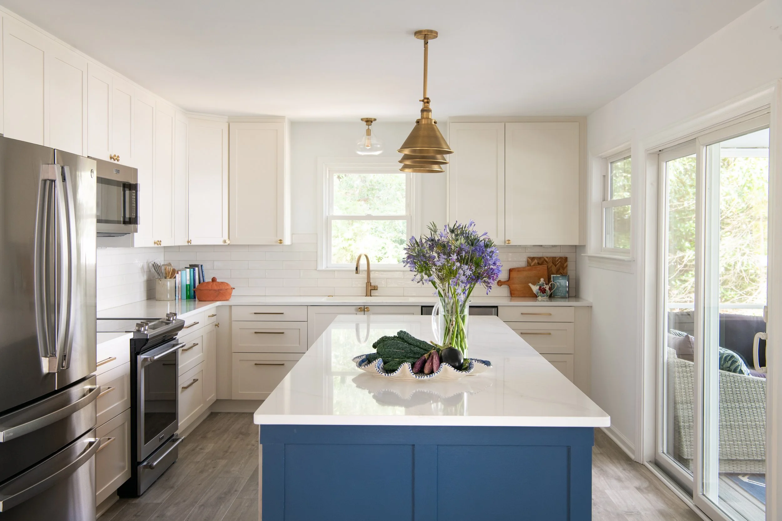 Bright kitchen with white cabinets, stainless steel appliances, a central island with a white countertop and blue base, a window behind the sink, and a bunch of purple flowers on the countertop.