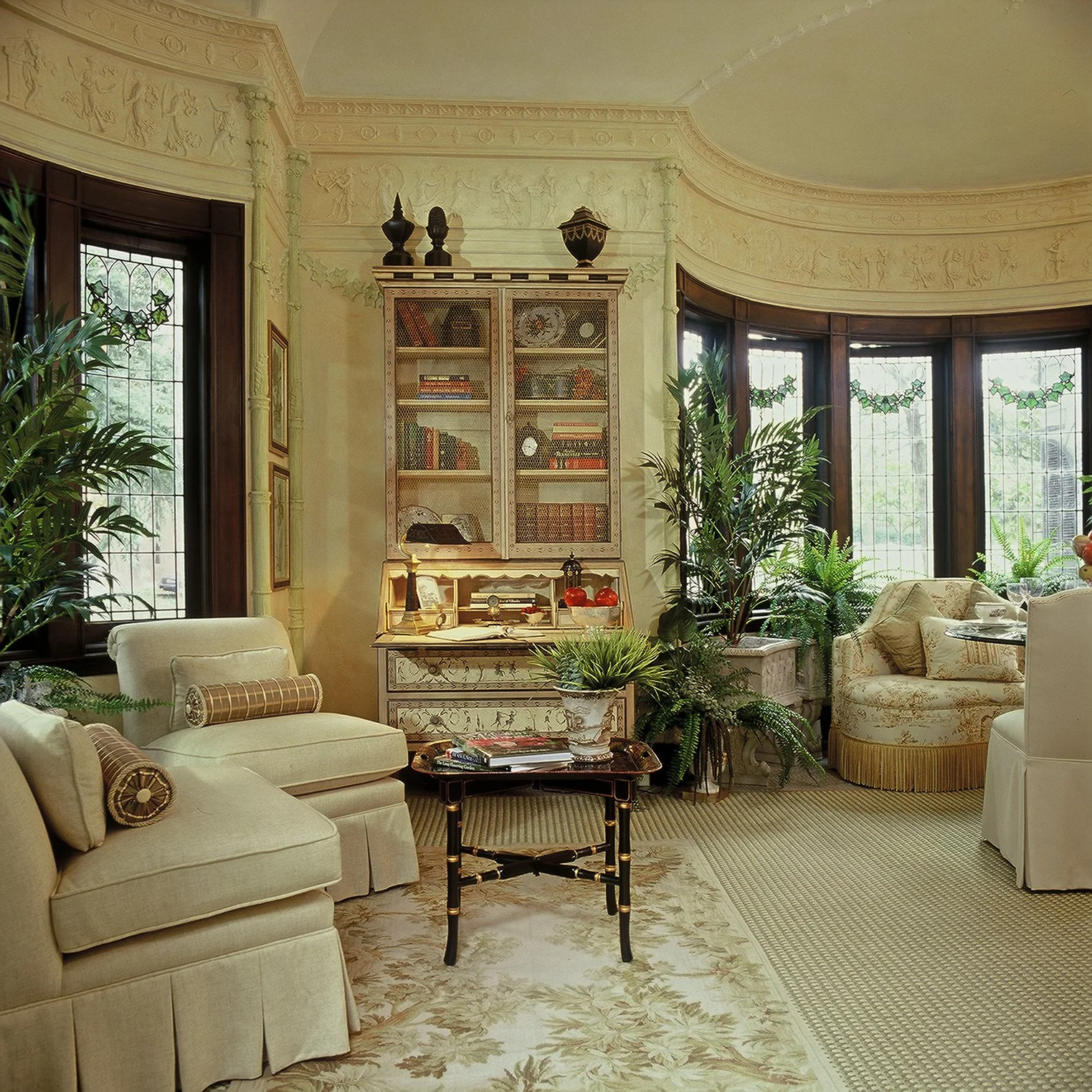 A cozy living room with cream-colored furniture, a black and gold coffee table, lush green plants, and a built-in bookshelf on the wall with window views of a garden outside.