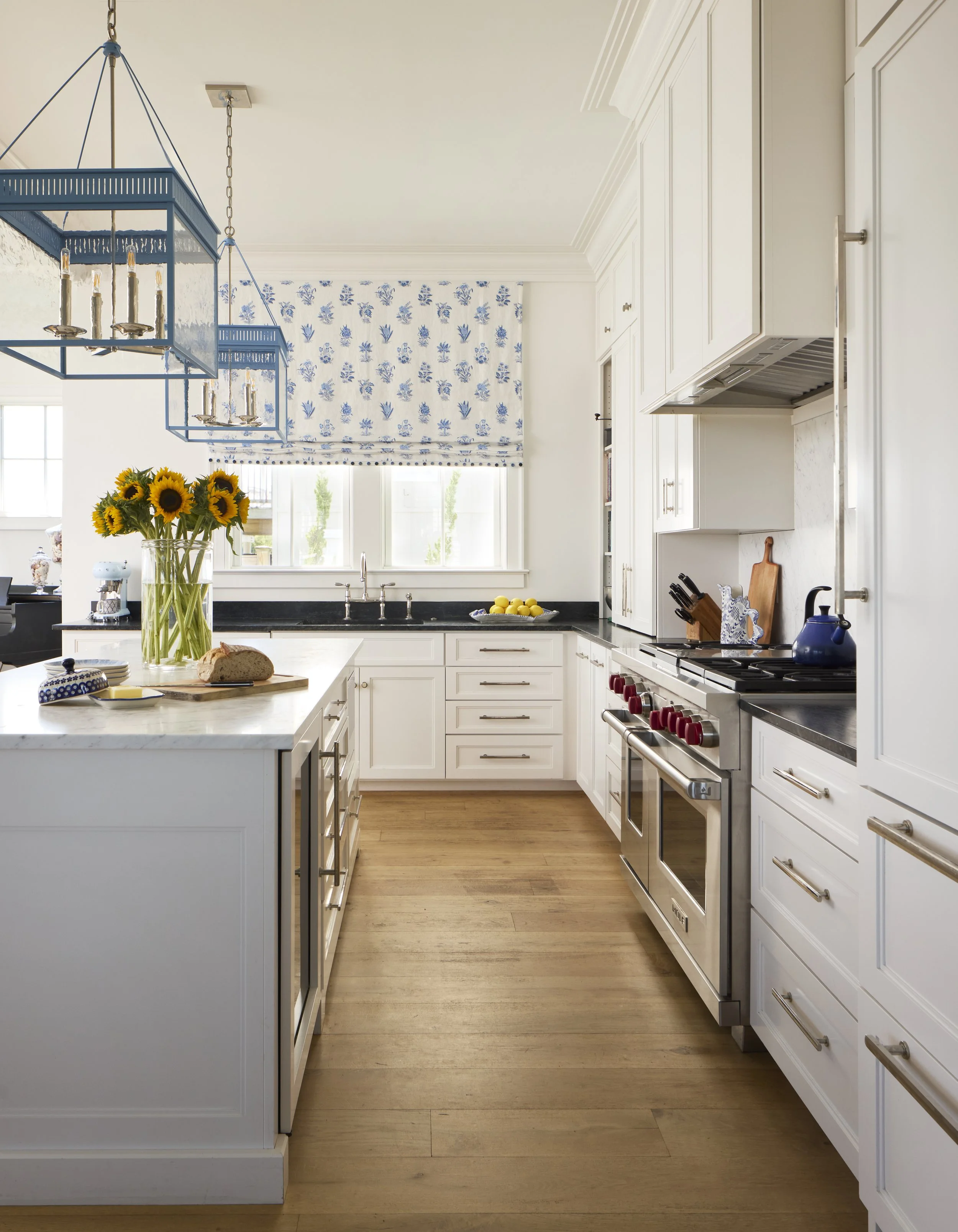 Bright kitchen with white cabinets, black countertops, and wooden flooring. A vase of sunflowers, a loaf of bread on a cutting board, and a bowl of lemons are on the island. Blue chandeliers are hanging from the ceiling. The window has a blue and white floral Roman shade.