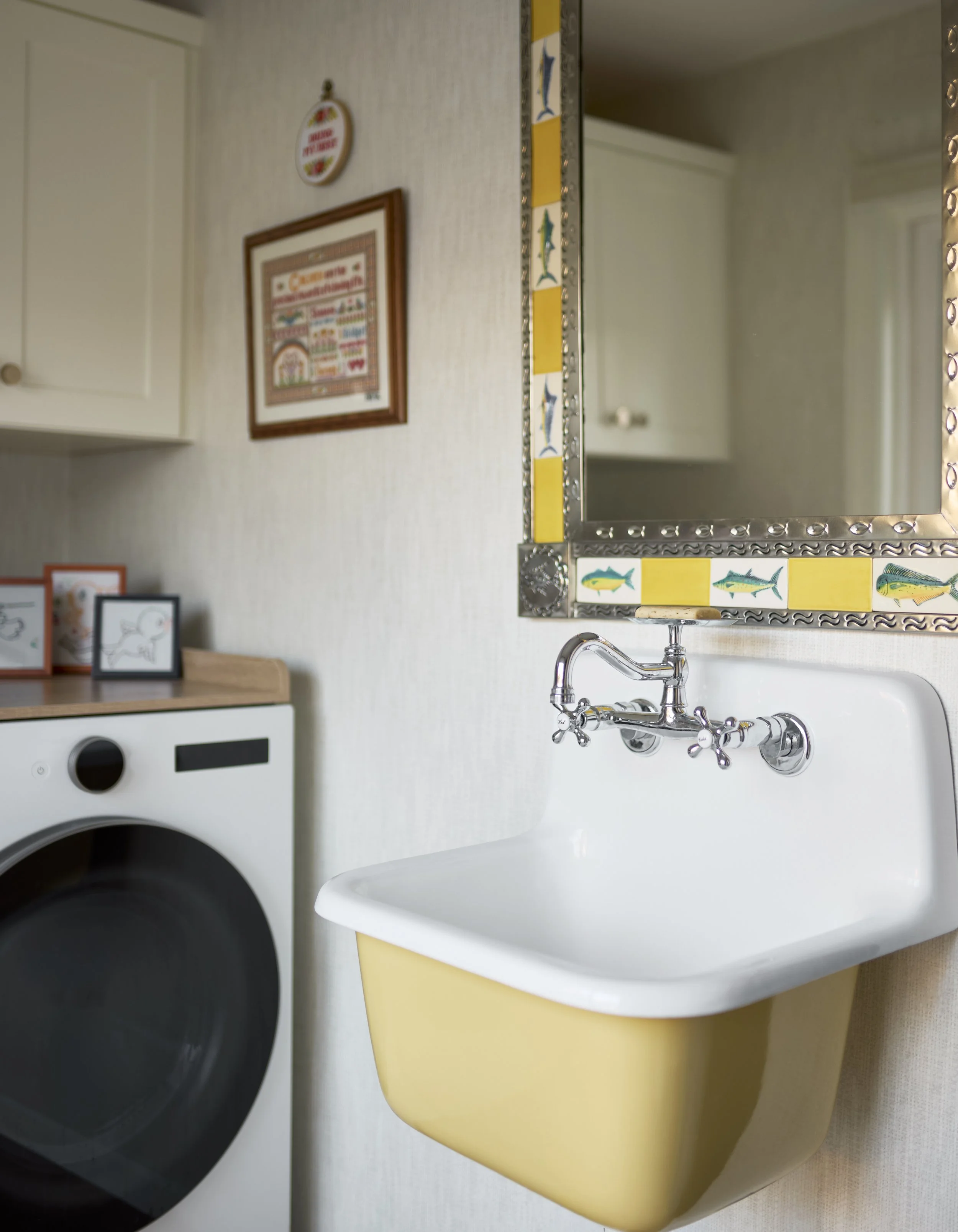 A laundry room with a white front-loading washing machine, a small yellow sink with a chrome faucet, a decorated mirror with fish tiles, and framed pictures on the wall.