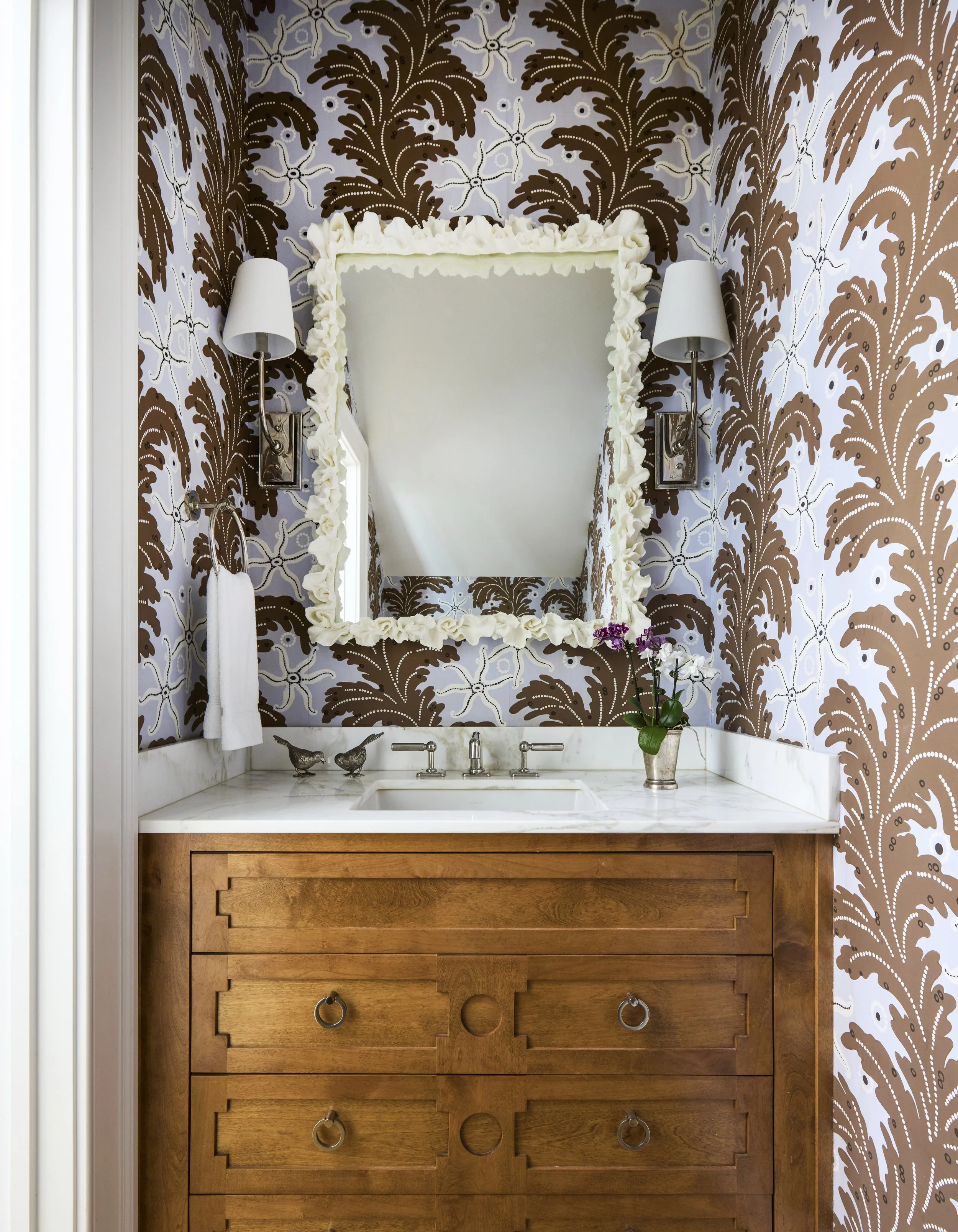 Powder room with starfish and acanthus wallpaper, mirror with coral like frame, two wall sconces, wooden dresser with marble countertop, small decorative bird figurines, potted orchid plant.